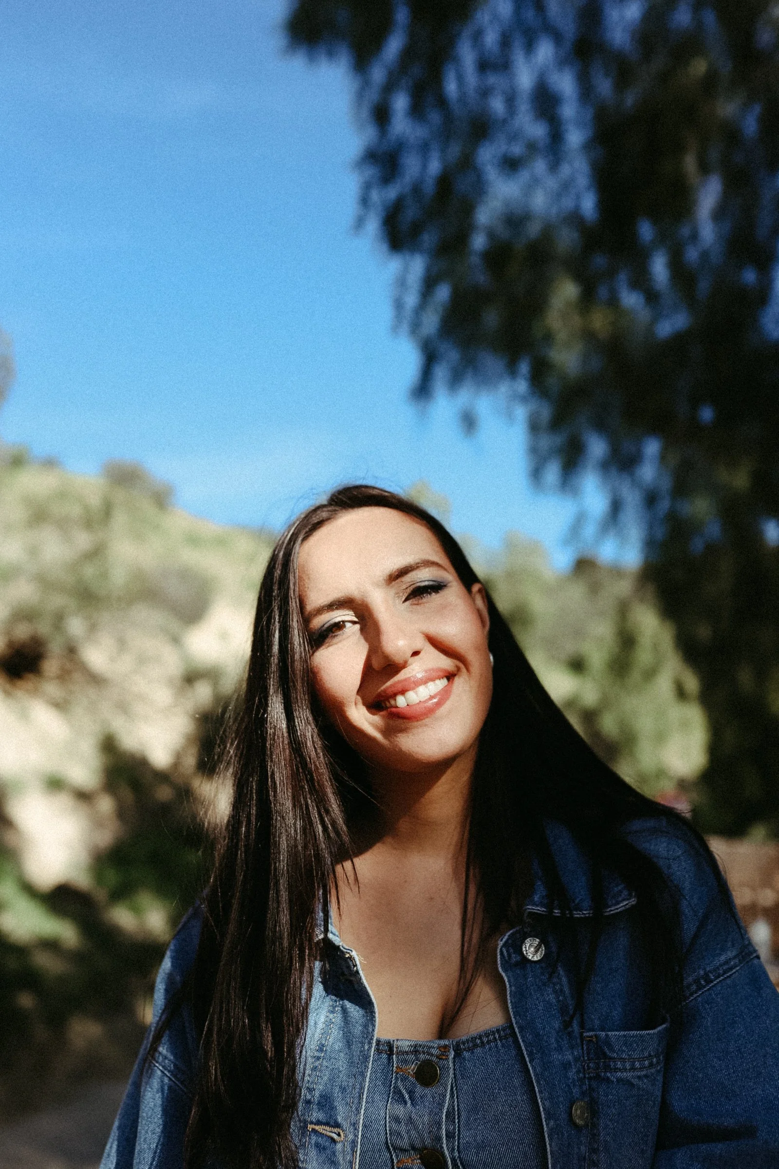Smiling young woman with long dark hair, wearing a denim jacket, outdoors with trees and blue sky in the background.
