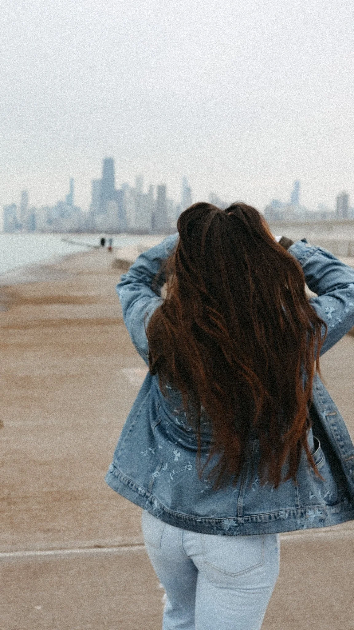 Woman with long brown hair wearing a blue denim jacket and white pants on a sandy beach with a city skyline in the background.