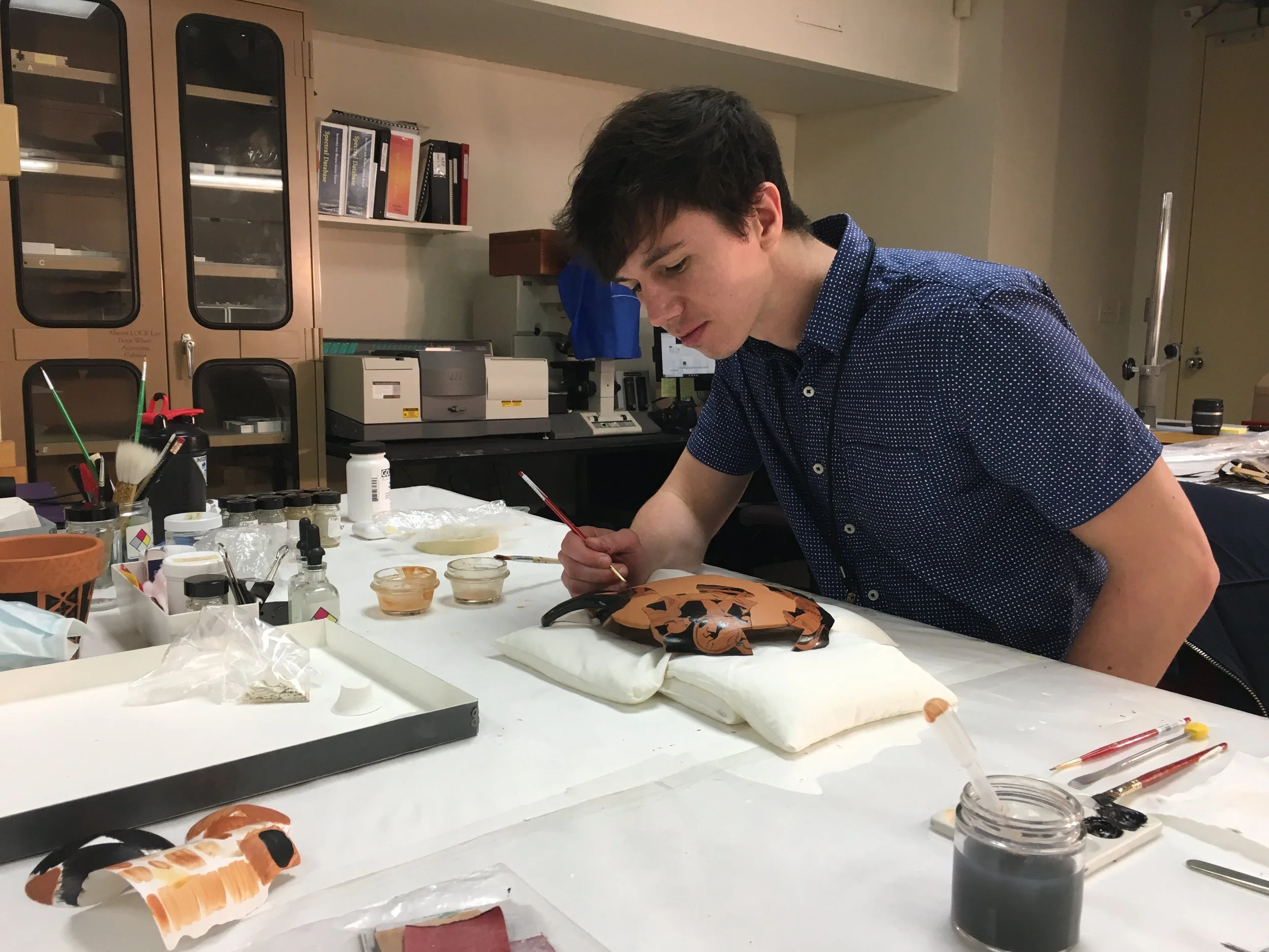A conservator restoring a Greek ceramic terracotta vessel in a studio, inpainting broken pottery. Studio specializing in objects conservation and ceramics in San Francisco, CA. Conservator / restorer of ceramics and art, objects and sculpture.