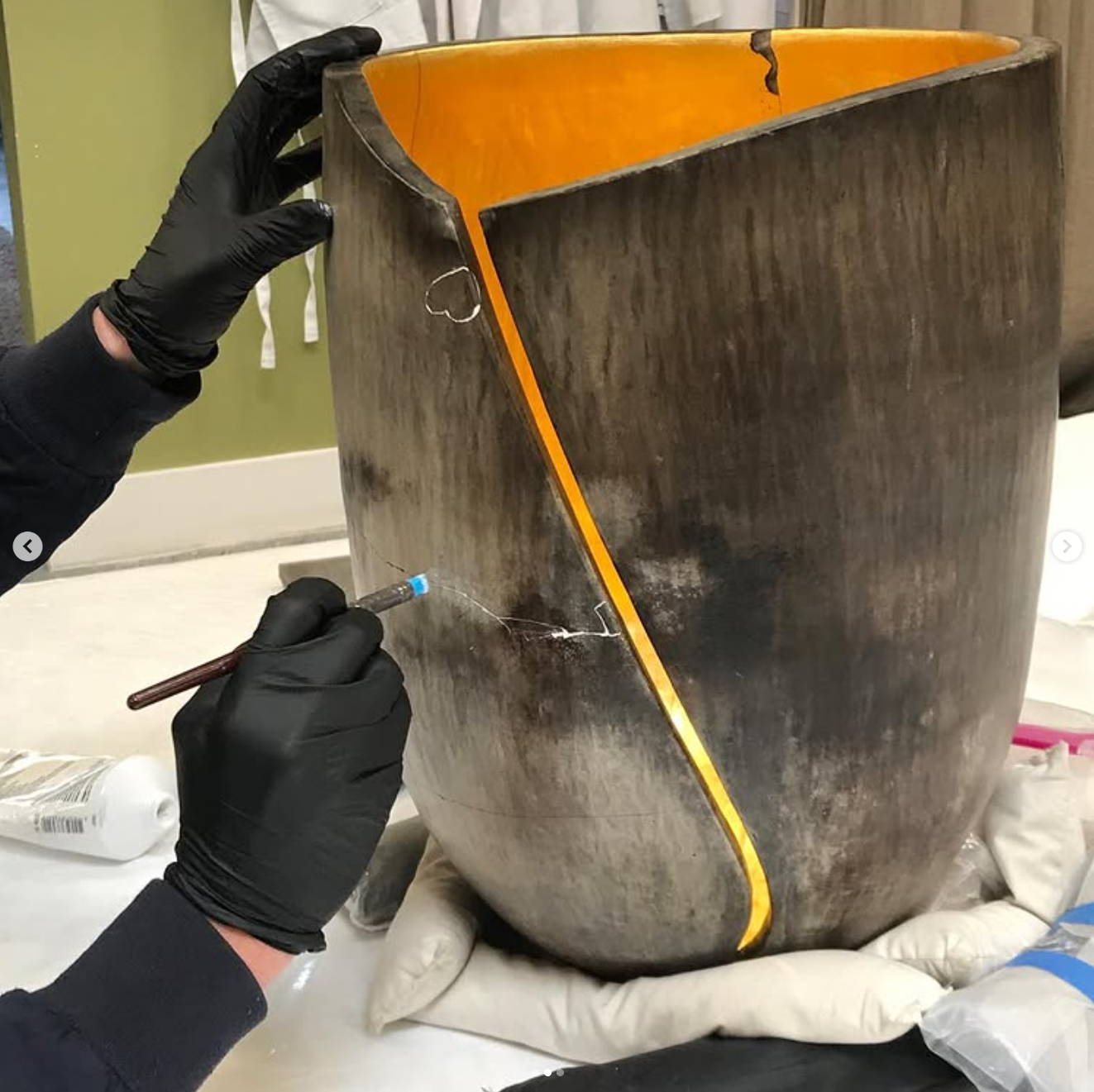 A person in black gloves is using a tool to carefully restore a large, ceramic vase with a gilded interior. The vase is supported on a cloth, in a conservation workshop. The conservator is filling losses on the exterior. 