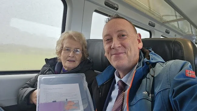 A man and an elderly woman sitting together on a train, with the man taking a selfie. The woman is holding a transparent folder with papers inside.