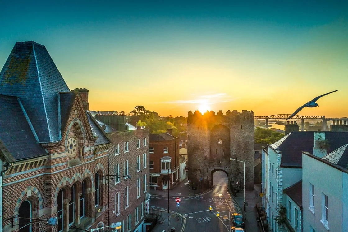 Sunset over a historic town with old brick buildings, a stone gate, and a bird flying.