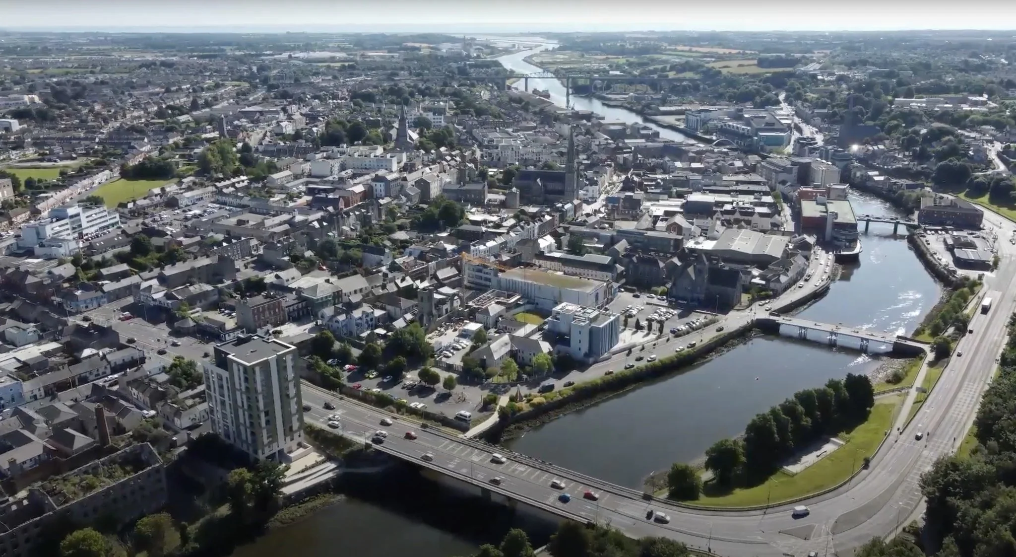 Aerial view of a city with a river running through it, showing bridges, roads, and various buildings, including a tall white high-rise.