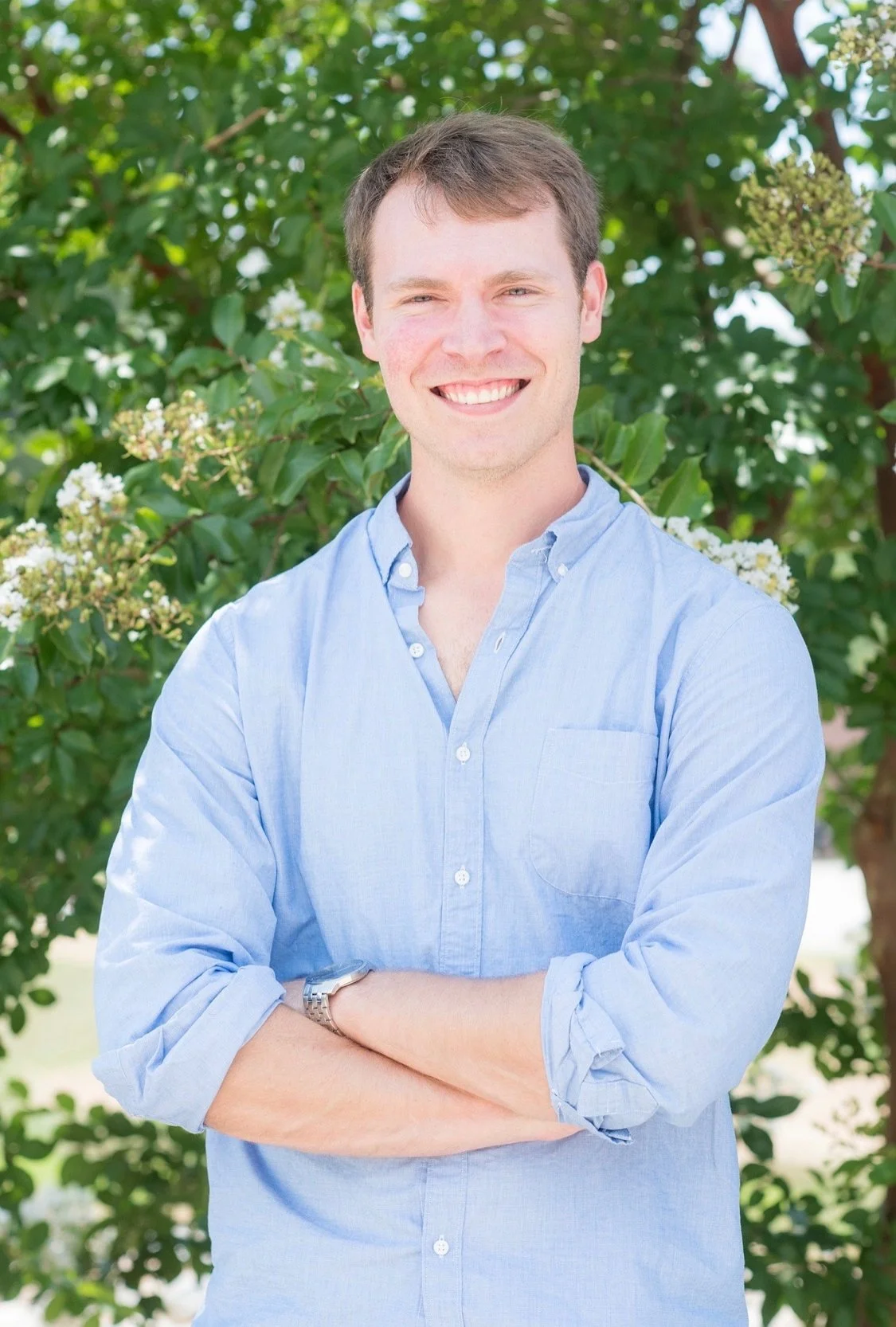 A young man with short brown hair, smiling, wearing a light blue button-up shirt with sleeves rolled up, standing outdoors with green leaves and white flowers in the background.
