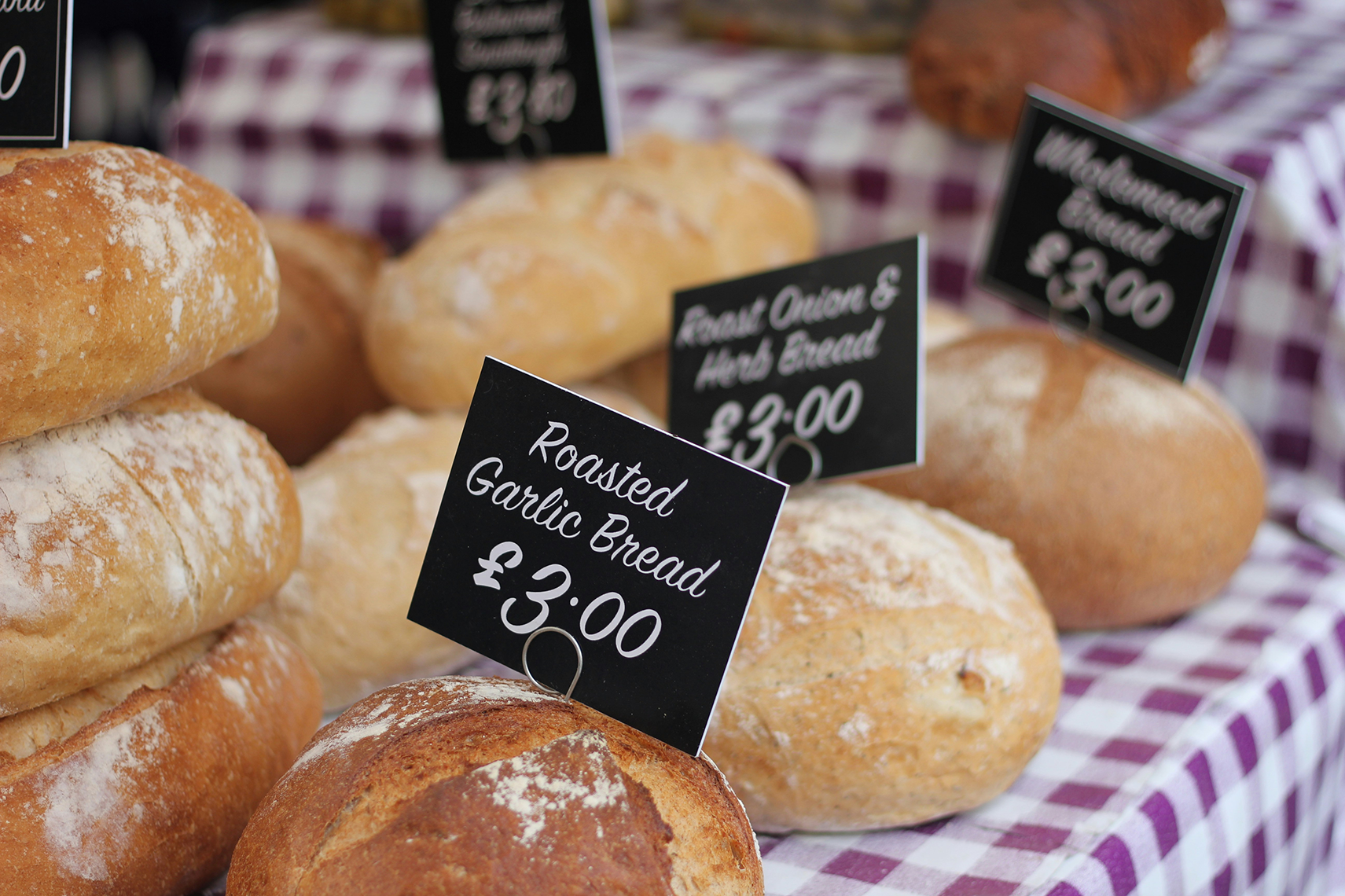 Assorted rustic bread loaves on a checkered tablecloth at a market stall with price tags.