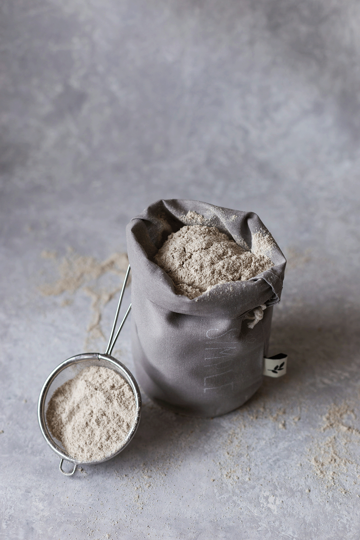 A gray cloth bag filled with flour, with a metal flour sifter and some spilled flour on a gray surface.