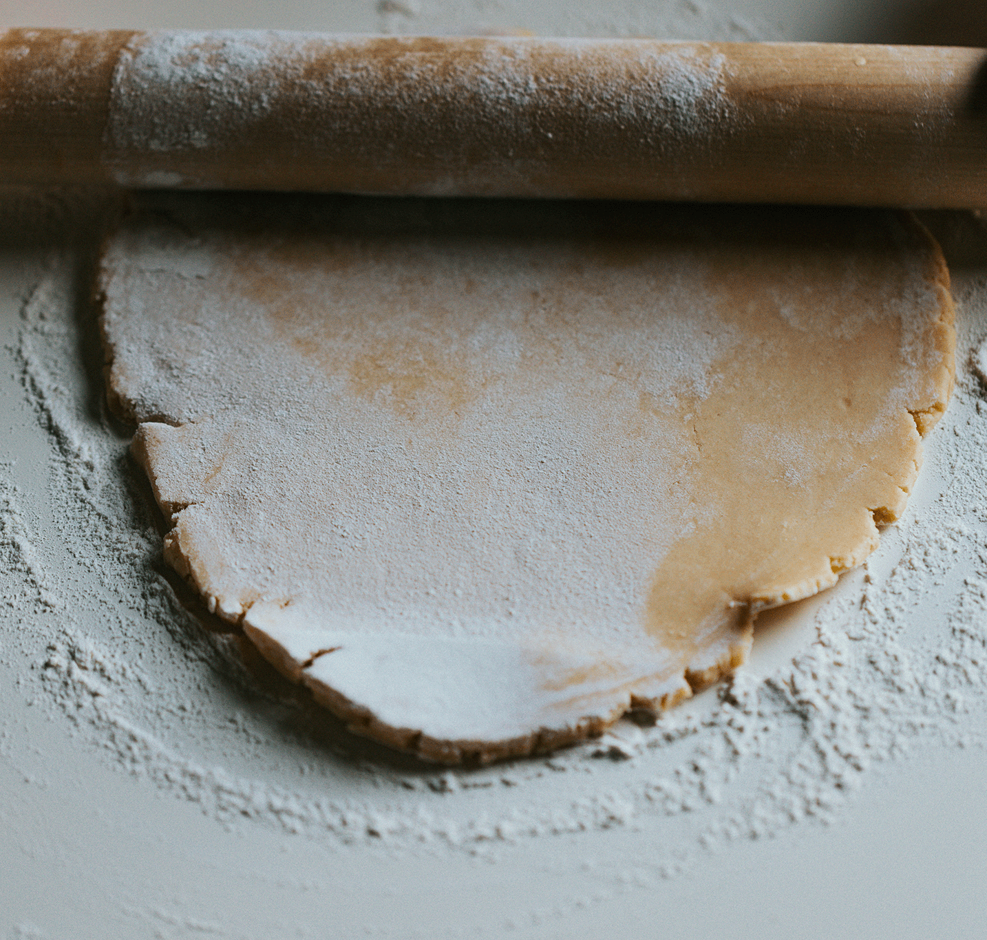 A rolling pin at the top with flour dusted on it, a sheet of dough sprinkled with flour underneath, and a portion of dough partially cut, dusted with flour, on a floured surface.