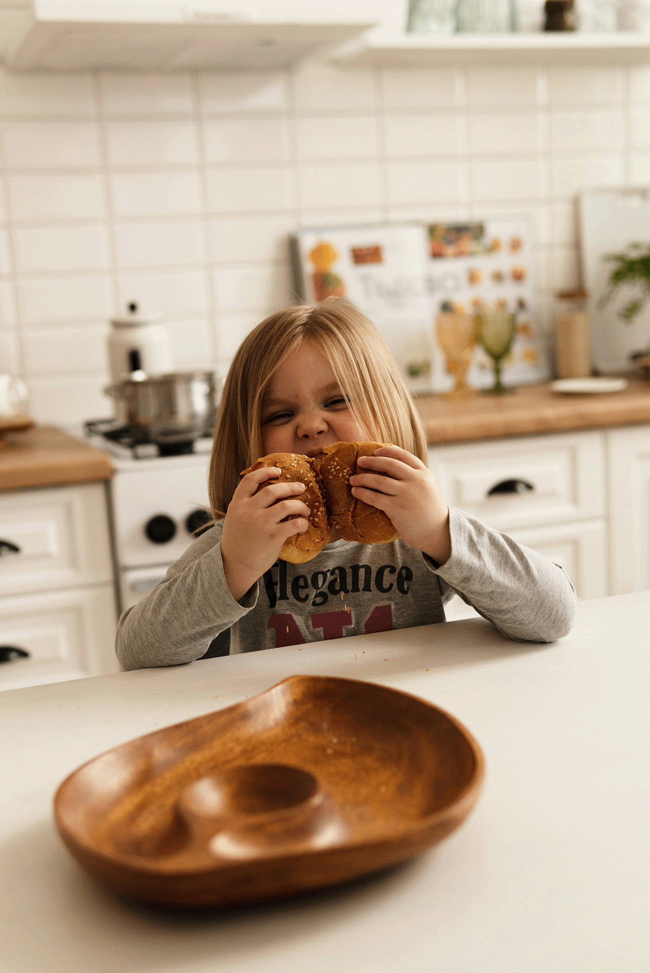 A young girl with red hair eating two sandwiches in a kitchen with a wooden countertop, white cabinets, and a stovetop in the background.