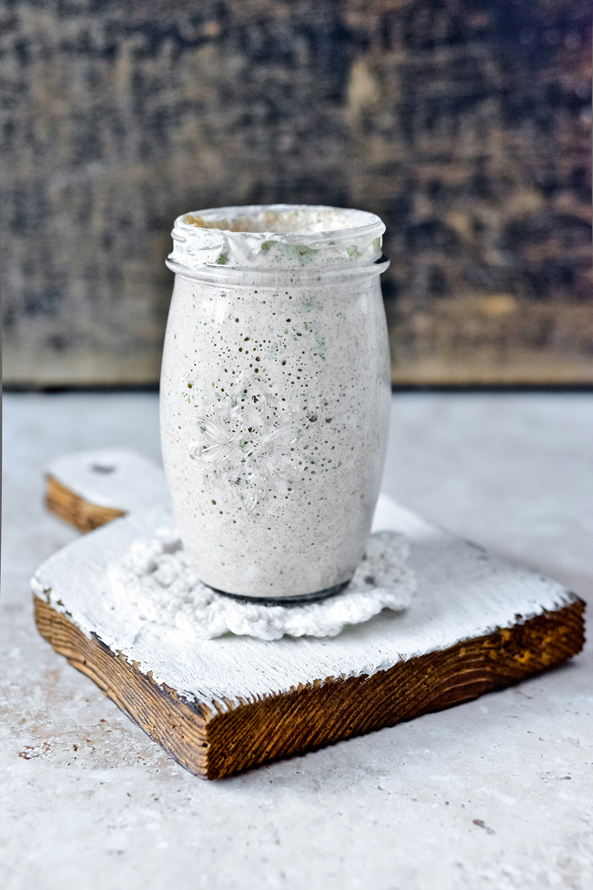 A glass jar filled with a white, speckled, snowy textured substance, garnished with whipped cream on top, placed on a rustic white wooden board with a piece of white cloth underneath, set against a dark, textured background.
