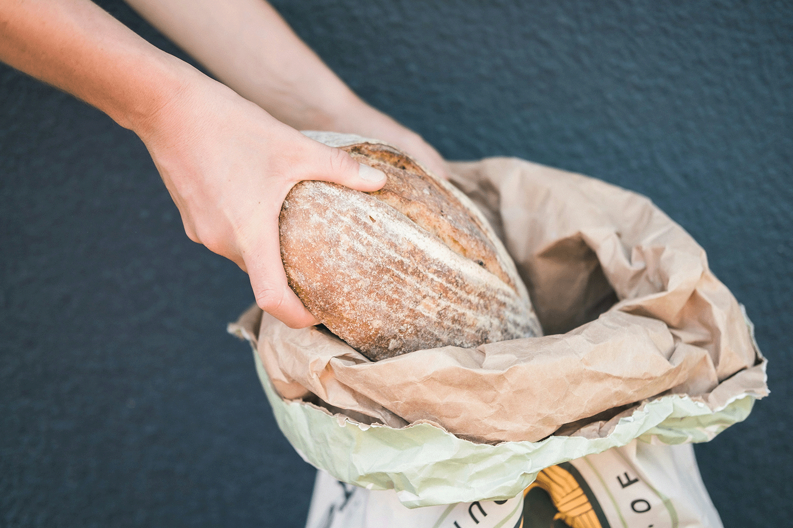 Person placing a loaf of crusty bread into a paper bag, with a dark surface underneath.