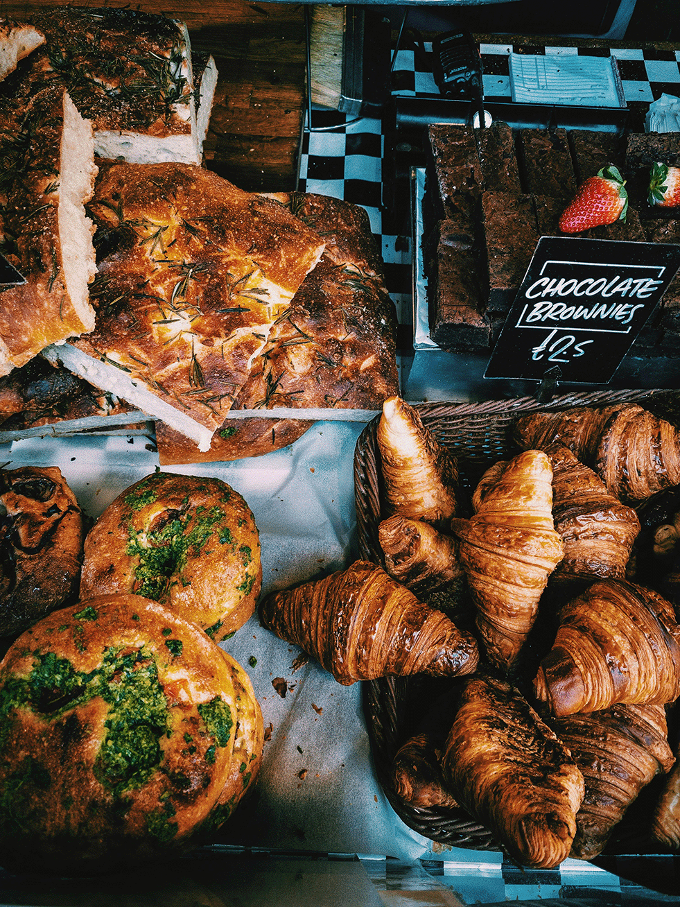 Assorted baked goods including croissants, bread, and muffins displayed on a table at a market, with a sign advertising chocolate brownies for $2.5.