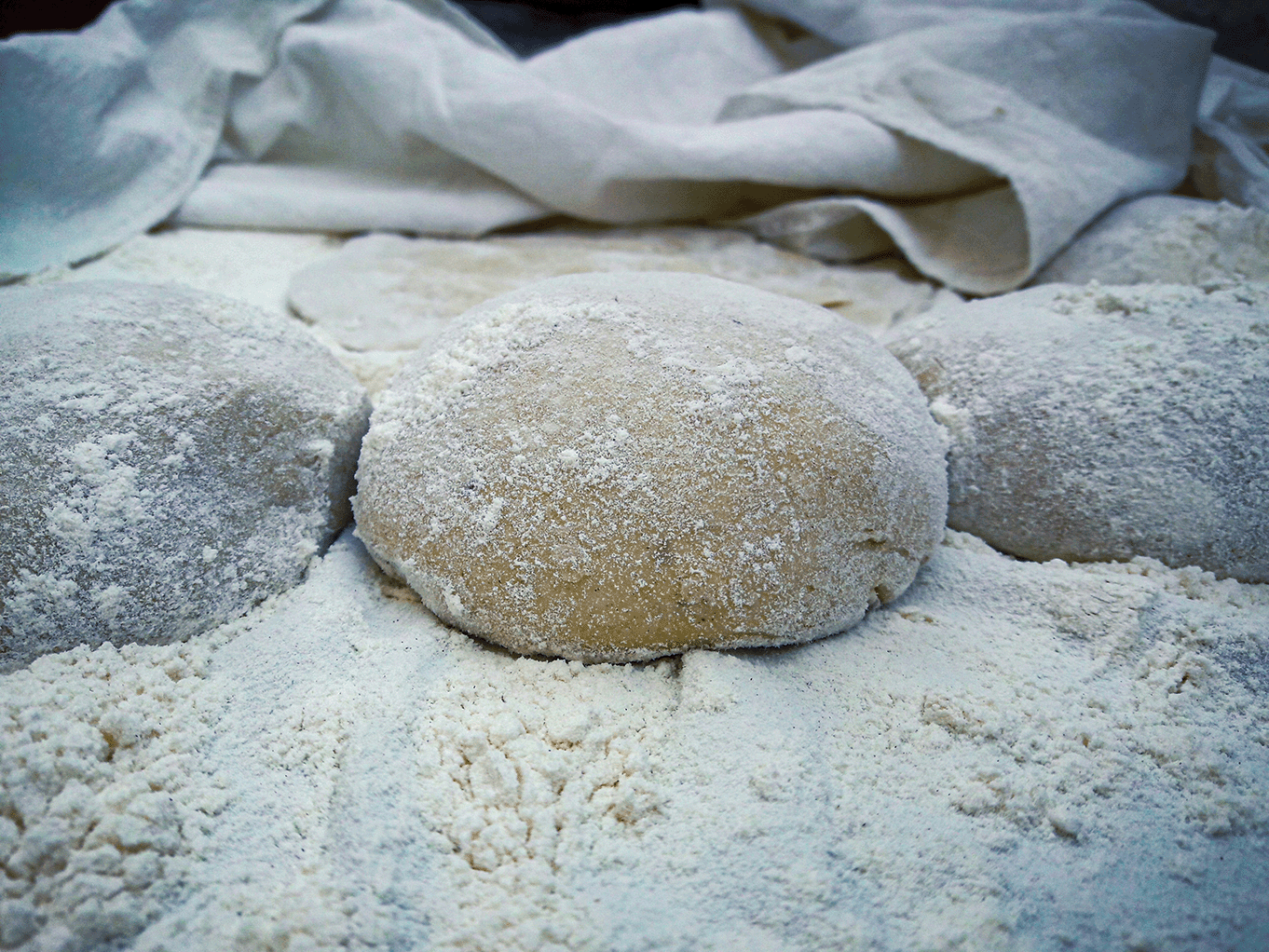 Close-up of three dough balls coated in flour, resting on a flour-dusted surface with a white cloth in the background.