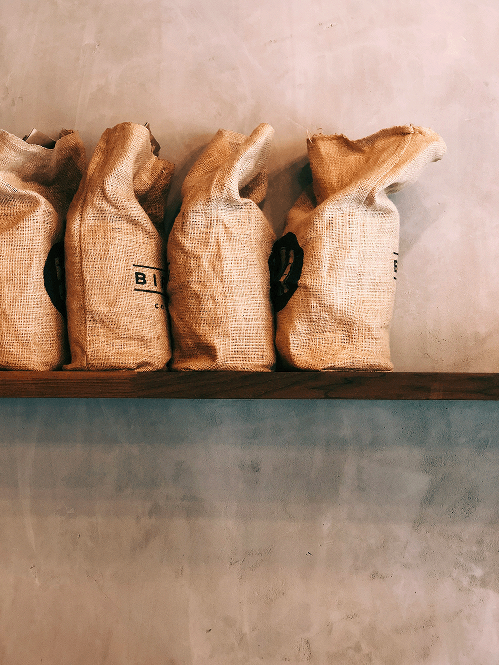 Four burlap coffee bean sacks lined up on a wooden shelf against a concrete wall.