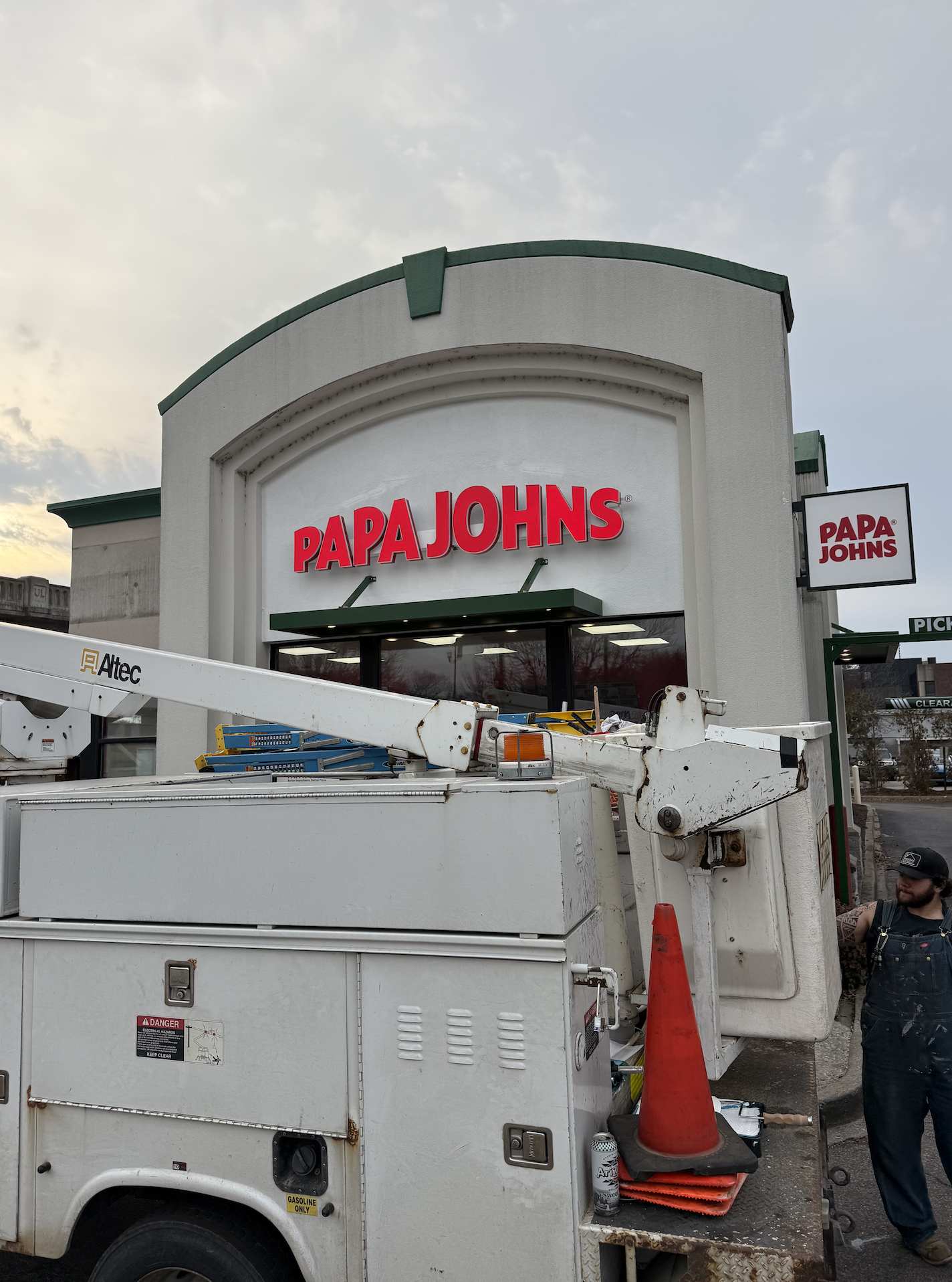 A Papa John's pizza restaurant exterior with a utility truck in front, a worker standing to the right, and an overcast sky.