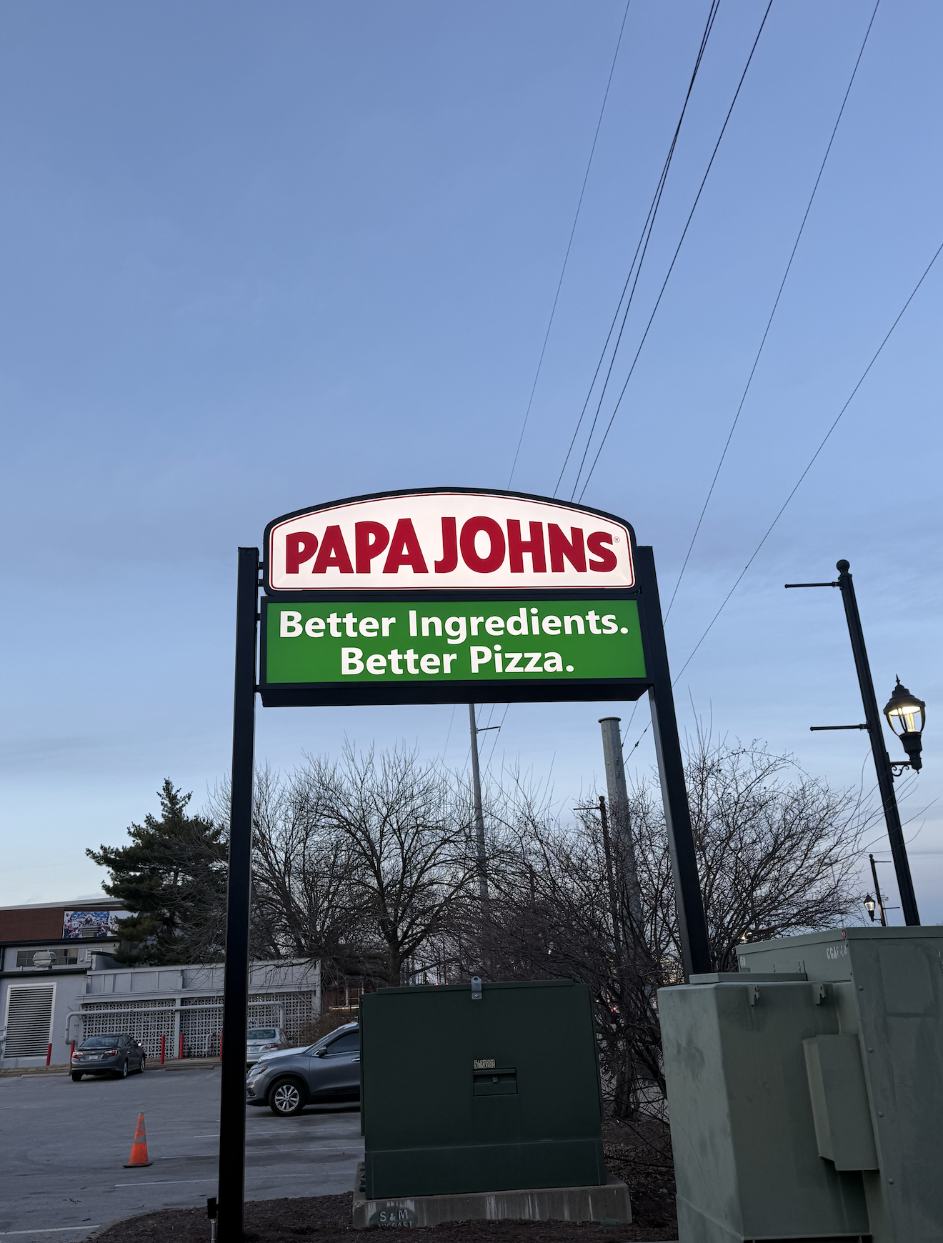 A Papa John's sign in an outdoor parking lot with a blue sky in the background, trees, cars, and street lamps nearby.