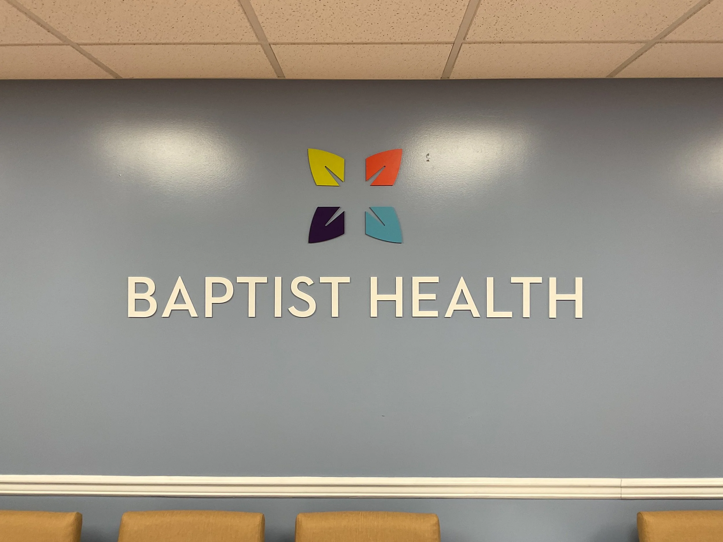 Baptist Health logo on a blue wall with four colorful leaves above the name. Four chairs are visible in the foreground.