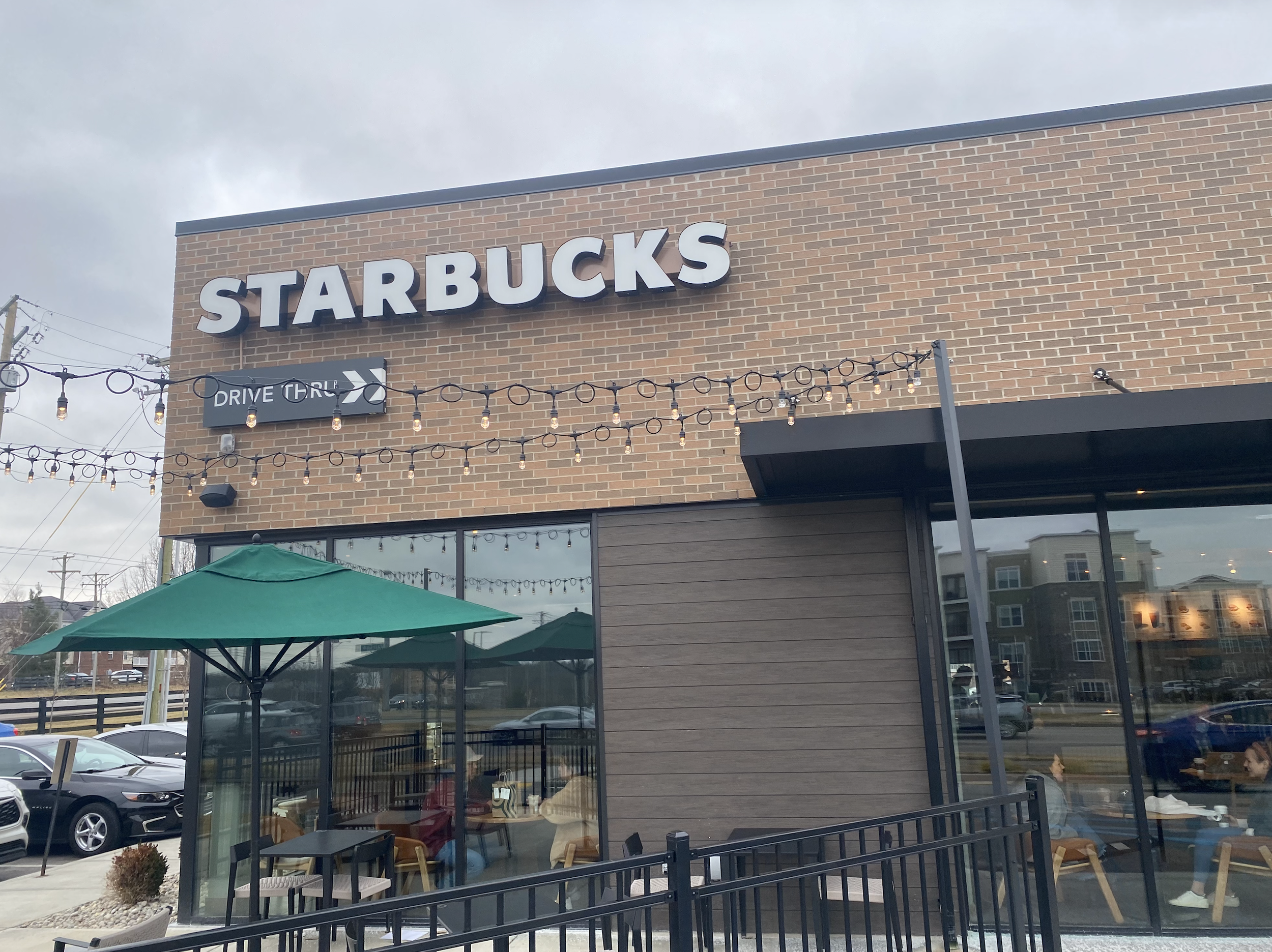 Exterior view of a Starbucks coffee shop with large logo sign, outside seating area with green umbrellas, and string lights hanging across the front.