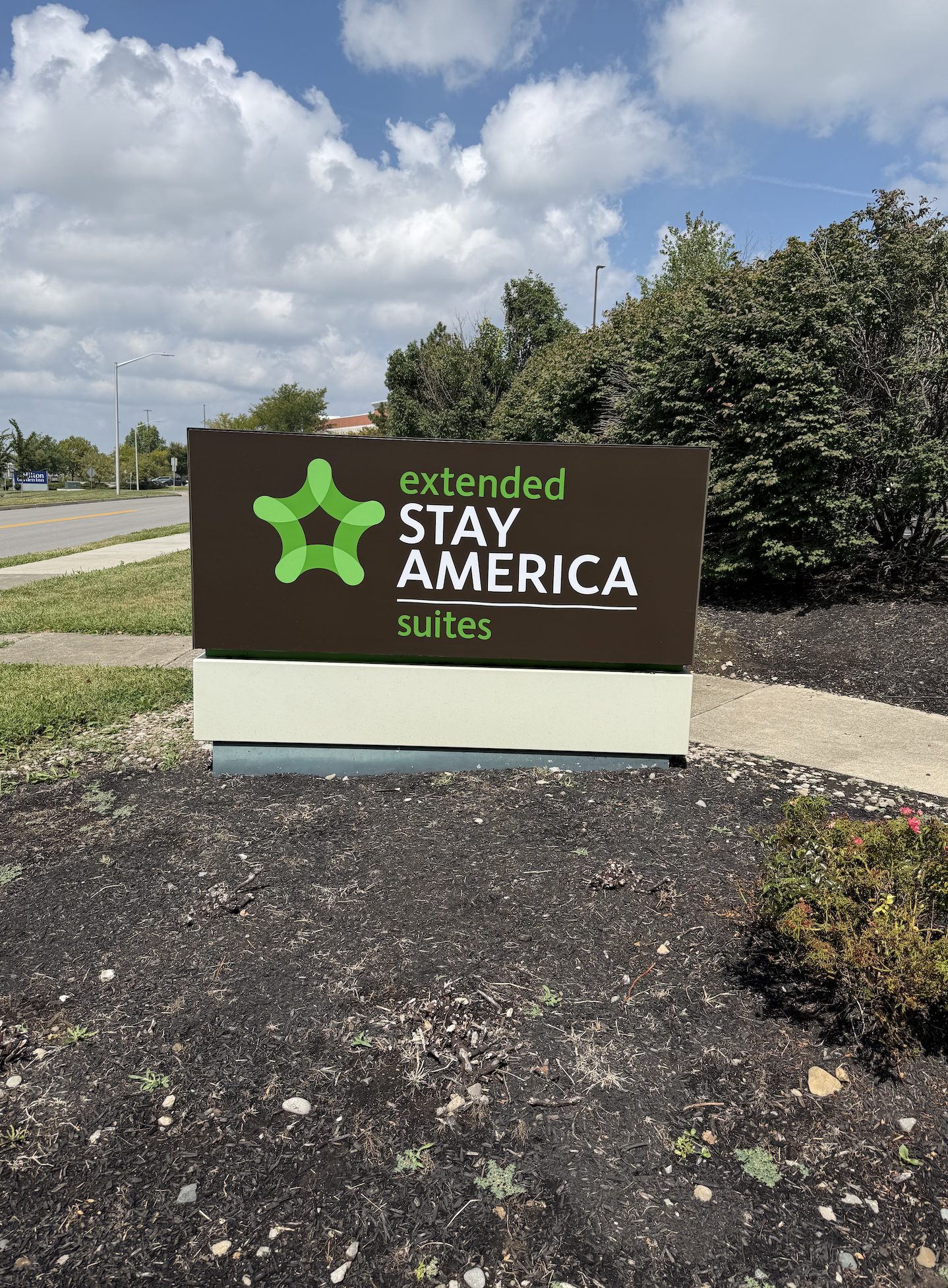 Sign for Extended Stay America Suites hotel with green abstract logo, on a sidewalk beside a dirt patch with small plants, under a partly cloudy sky.