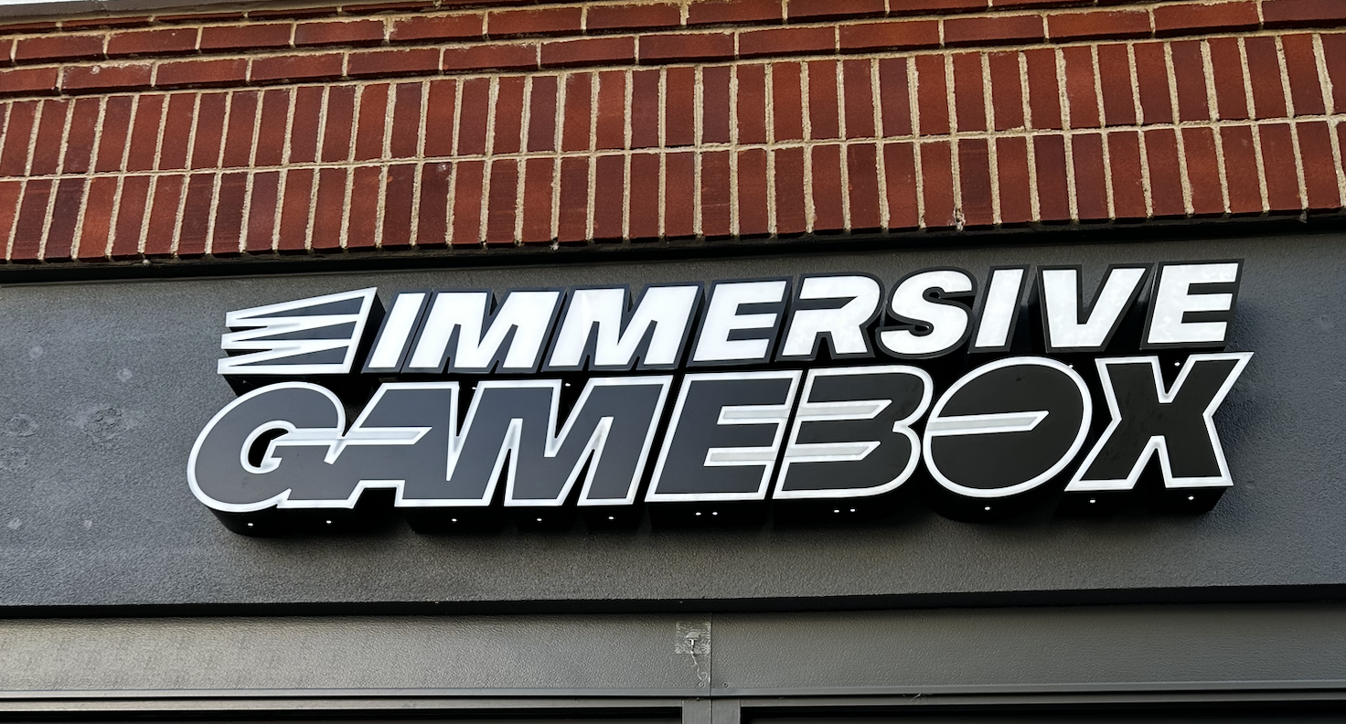 Illuminated sign with the words "Immersive Gamework" in bold, stylized letters, mounted on a wall with a brick and gray surface background.