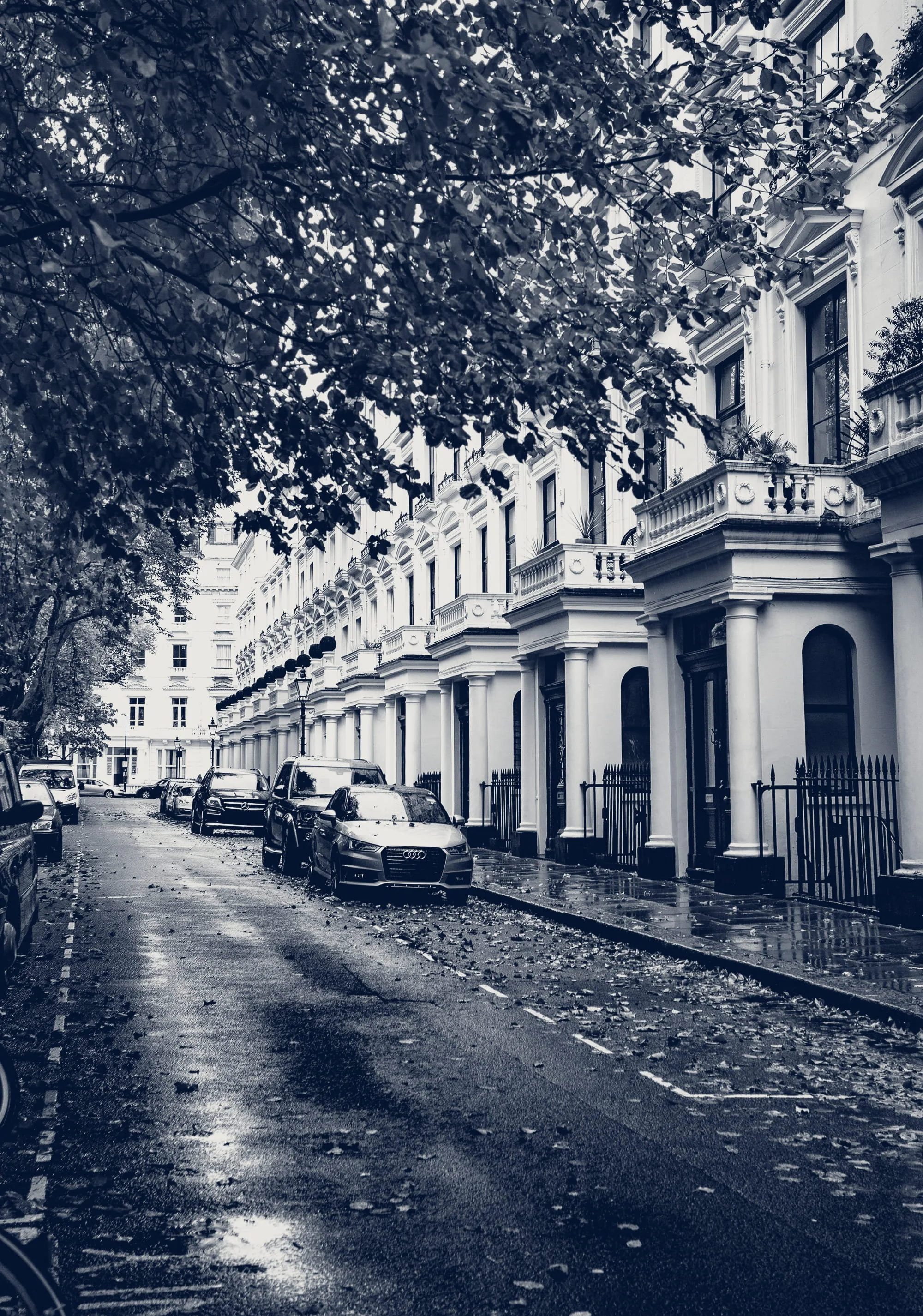 Street view in front of European-style white building with arches and balconies, parked cars, wet pavement, and overhanging trees, black and white photo.