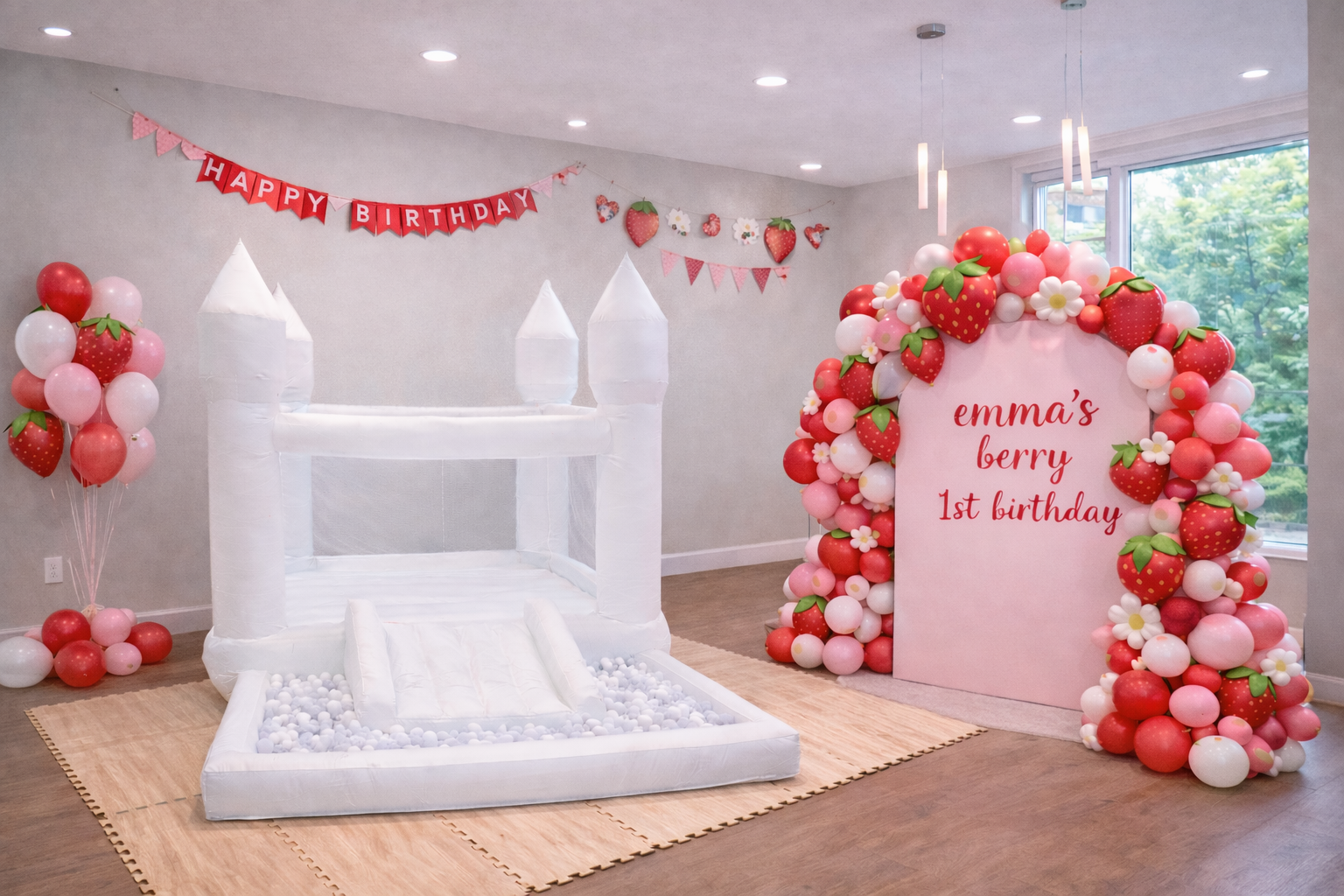Indoor birthday party setup with a white inflatable slide, a strawberry-themed balloon arch, and a pink backdrop reading 'emma's berry 1st birthday,' decorated with strawberry and flower balloons.