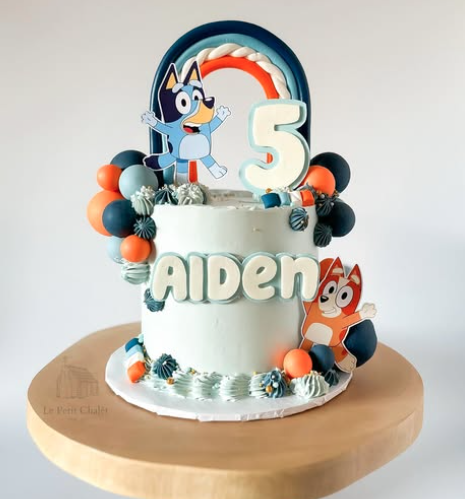 Two-tiered birthday cake decorated with Paw Patrol characters, a rainbow topper, a large number 5, and the name 'Aiden', placed on a wooden cake stand.