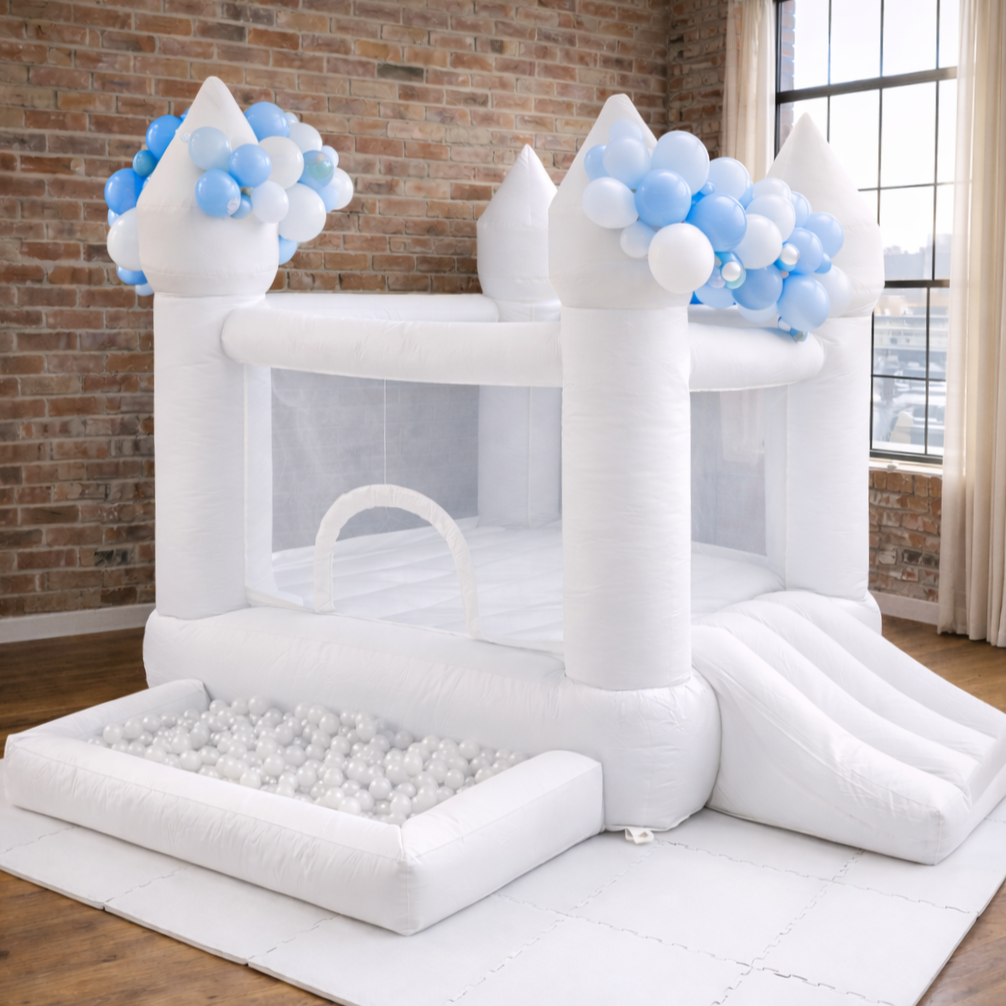White inflatable castle with blue and white balloons decoration, a slide, and a ball pit, set up indoors near a window with curtains.
