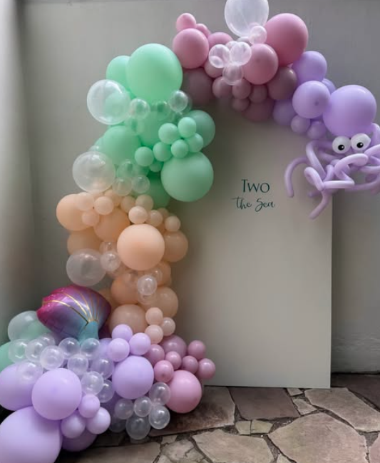Colorful balloon arrangement with pastel balloons in green, pink, purple, peach, and clear, shaped into an arch with a purple octopus balloon on the right, placed next to a white board with text 'TWO the Sea'.
