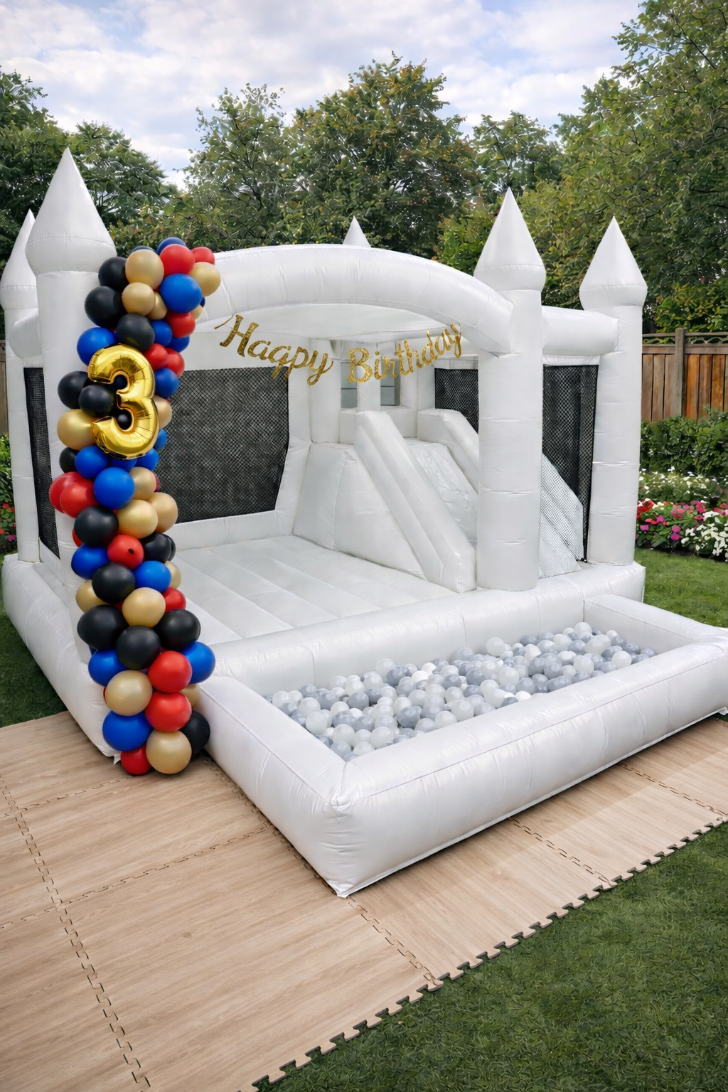 Inflatable white castle bounce house with a slide, decorated with a gold 'Happy Birthday' banner, and a column of red, black, gold, and blue balloons with a gold number 3 balloon, set up outdoors on a grassy area and wooden floor mats.