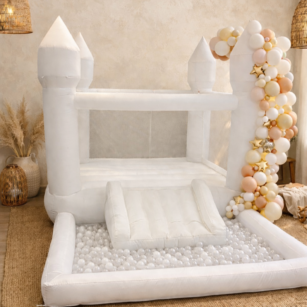 A white inflatable castle with a ball pit, balloon garland, and a small slide, decorated for a celebration or party.