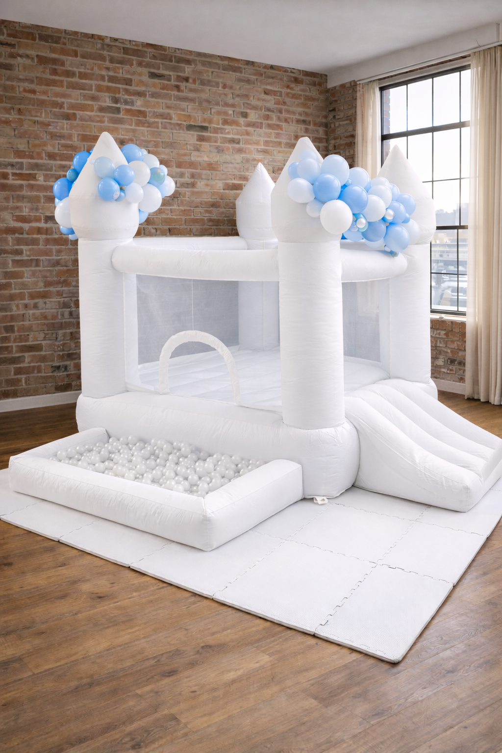 White inflatable castle with blue and white balloons, a slide, and a ball pit, set up indoors on a white padded mat in a room with brick wall and large window.