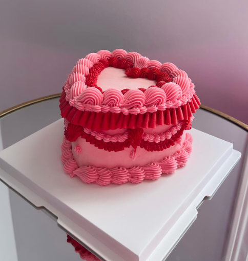 Heart-shaped pink and red layered cake with decorative frosting, placed on a white cake board.