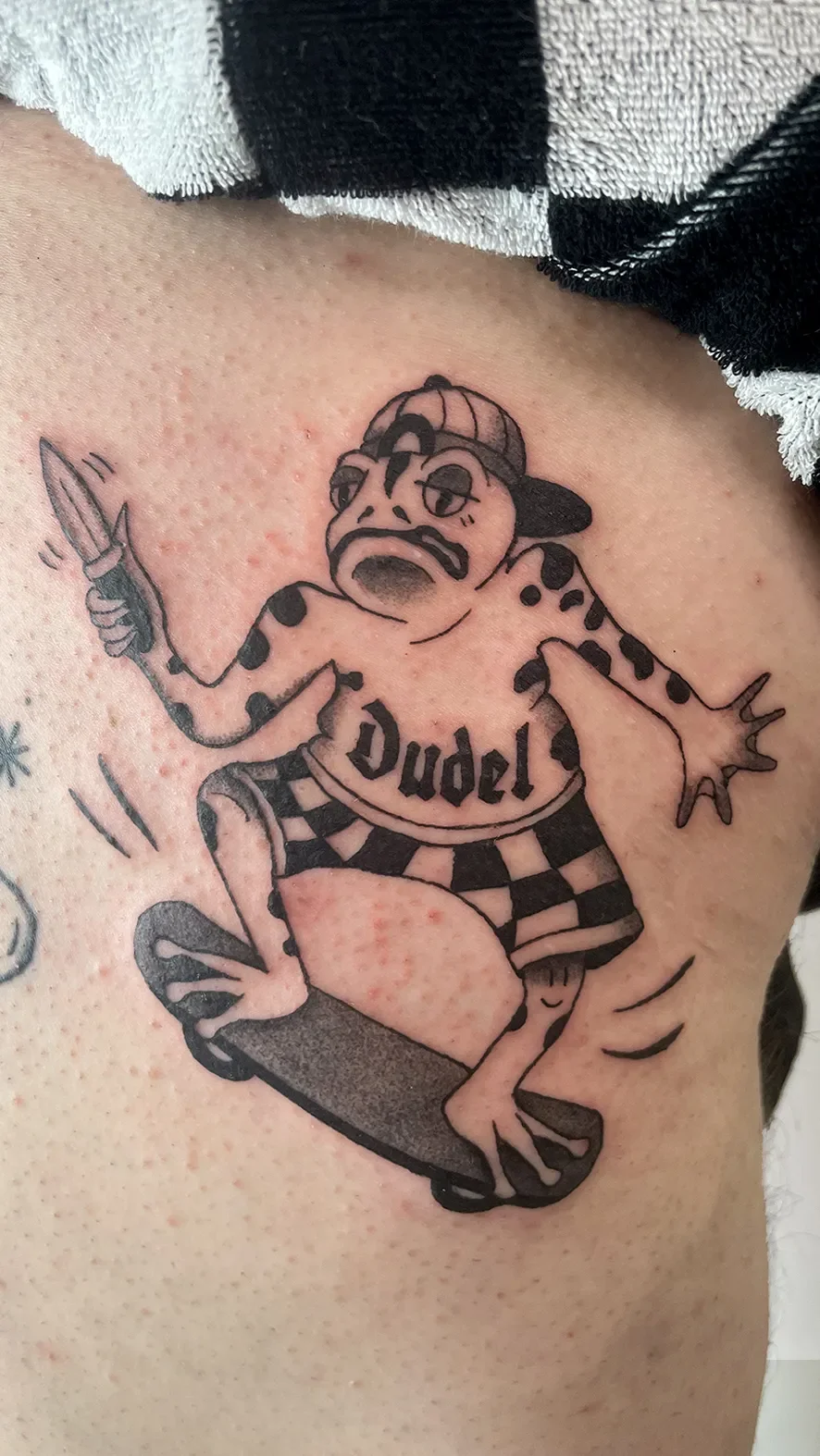 Tattoo eines humanoiden Tieres mit einem Skateboard, trägt eine Mütze und ein T-Shirt mit der Aufschrift "Dudel".