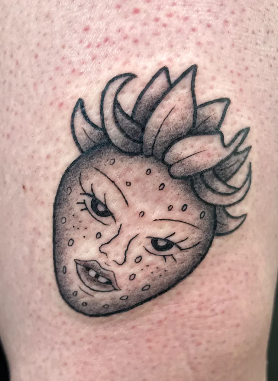 Tattoo eines stilisierten Erdbeers mit einem menschlichen Gesicht und einem Pflänzchen auf der Spitze, das eine Mischung aus Frucht und menschlichem Gesicht darstellt.