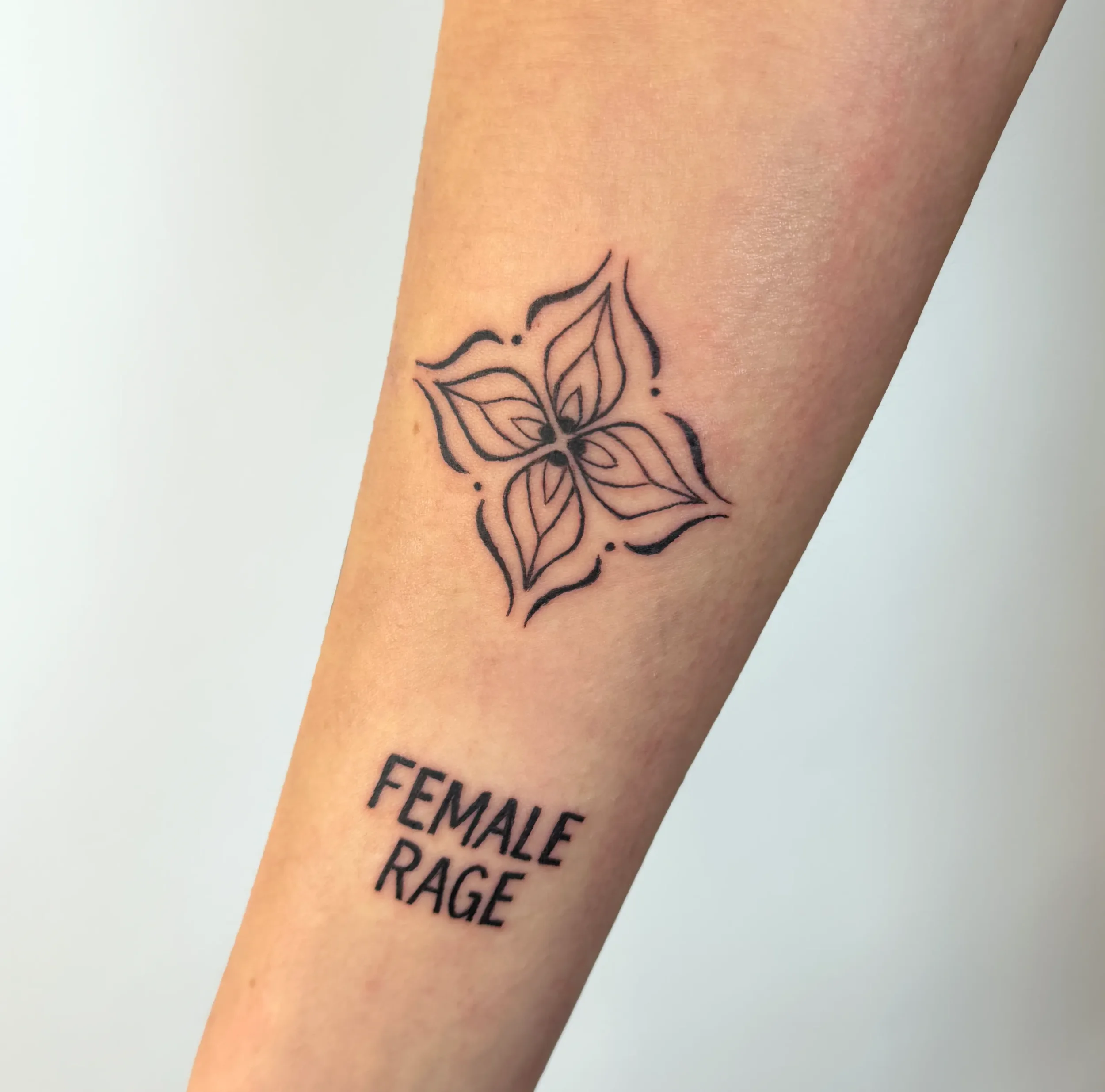 Tattoo eines schwarzen, stilisierten Schmetterlings mit den Worten 'Feminine Rage' darunter auf der Haut.