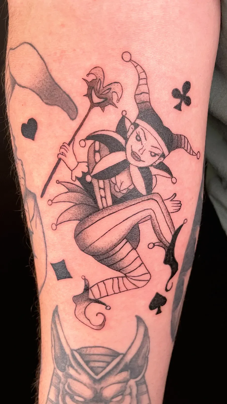 Tattoo einer lachenden Harlequin-Figur im Karnevalskostüm mit Hörnern, Halsschmuck, Zauberstab und fliegenden Spielkartensymbolen, auf einem Arm.