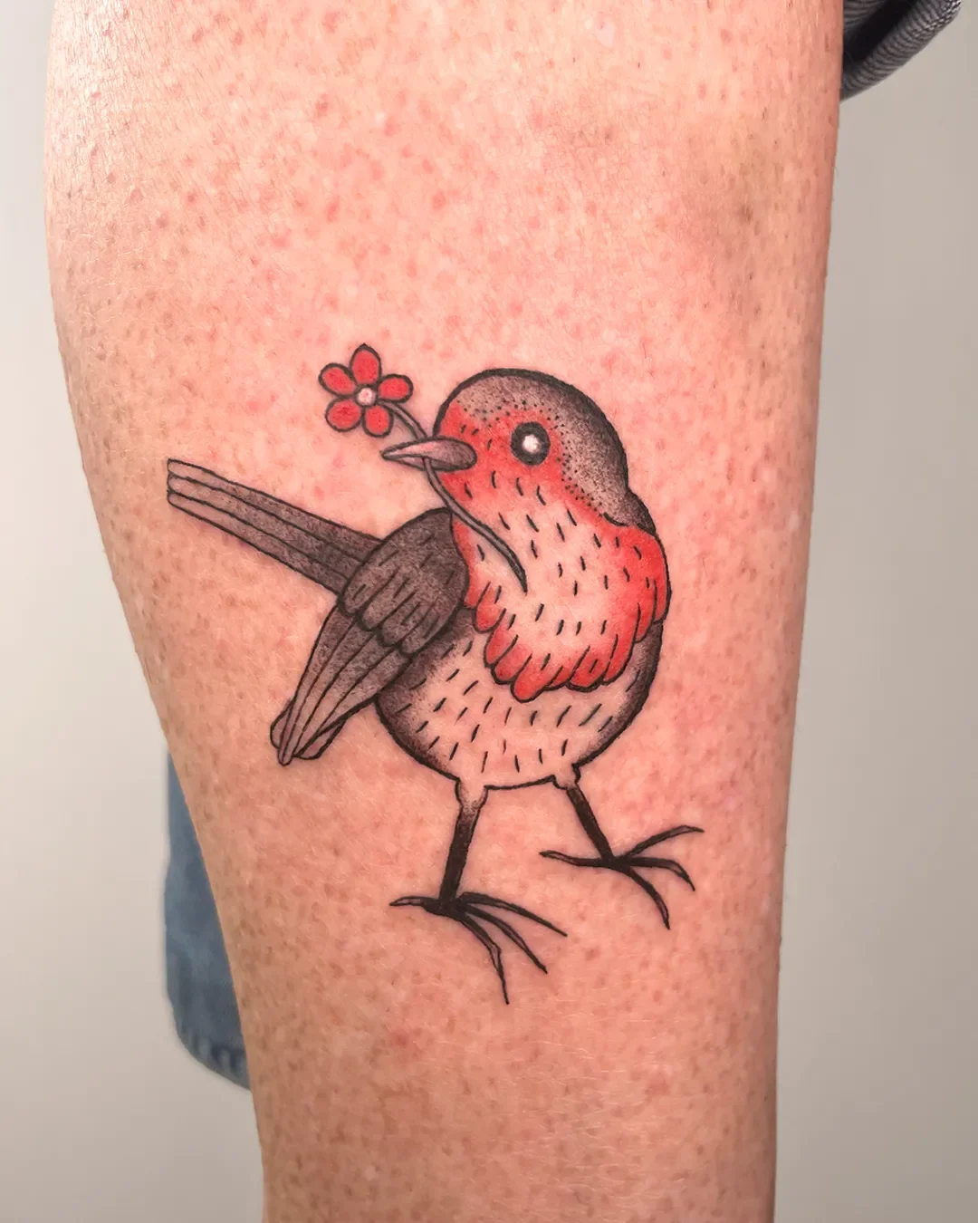Tattoo eines bunten Vogel mit roter Brust, der ein kleines rotes Blumen in den Schnabel hält.