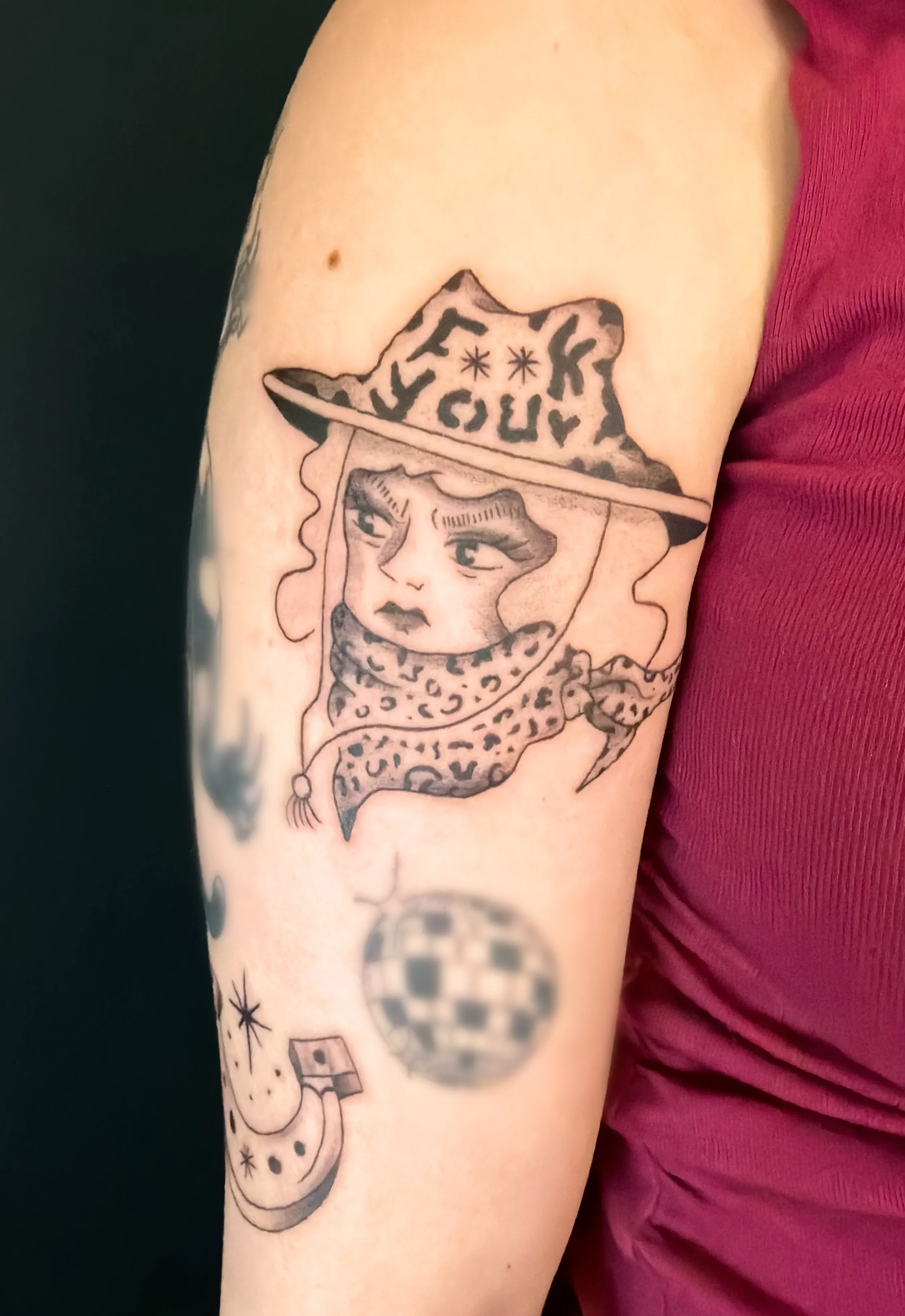Tattoo eines Mädchengesichts mit Hut und Schleife, umgeben von ungewöhnlichen Symbolen auf dem Oberarm.