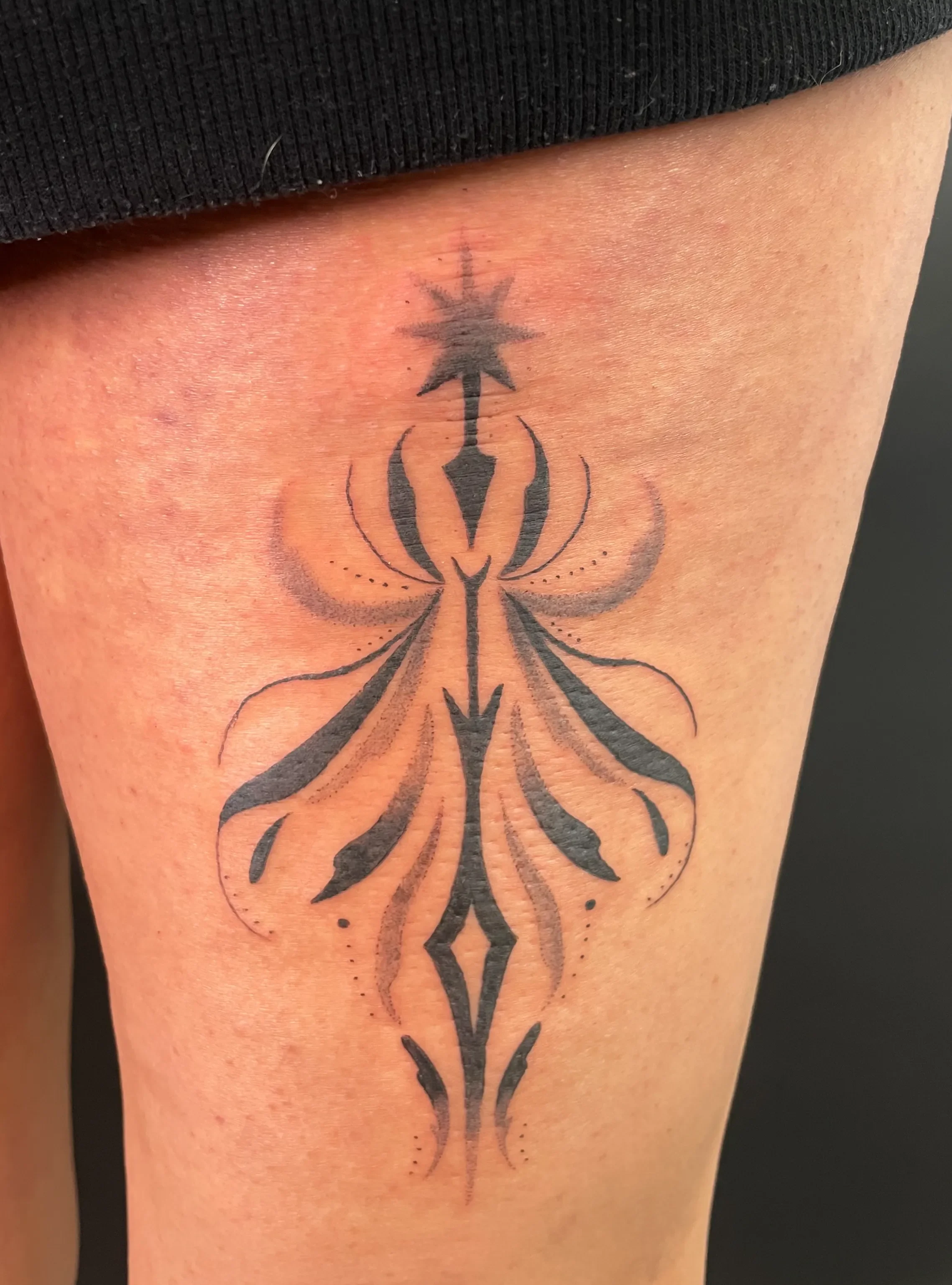 Schwarze Tribal-Tattoo auf einer Haut an Oberschenkel, bestehend aus geometrischen und symbolischen Mustern, inklusive Stern- und Pfeilsymbolen.