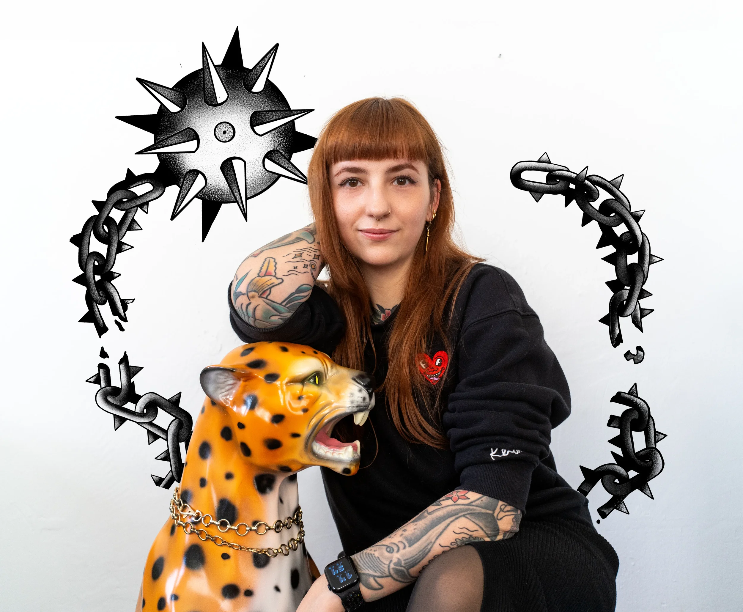 Junge Frau mit roten Haaren, Tätowierungen, schwarzem Pullover mit Herzaufnäher, sitzt neben einer leuchtend gefärbten Leoparden-Statue mit Kette, vor einem weißen Hintergrund, umgeben von schwarzer Ketten- und Stachel-Dekoration.