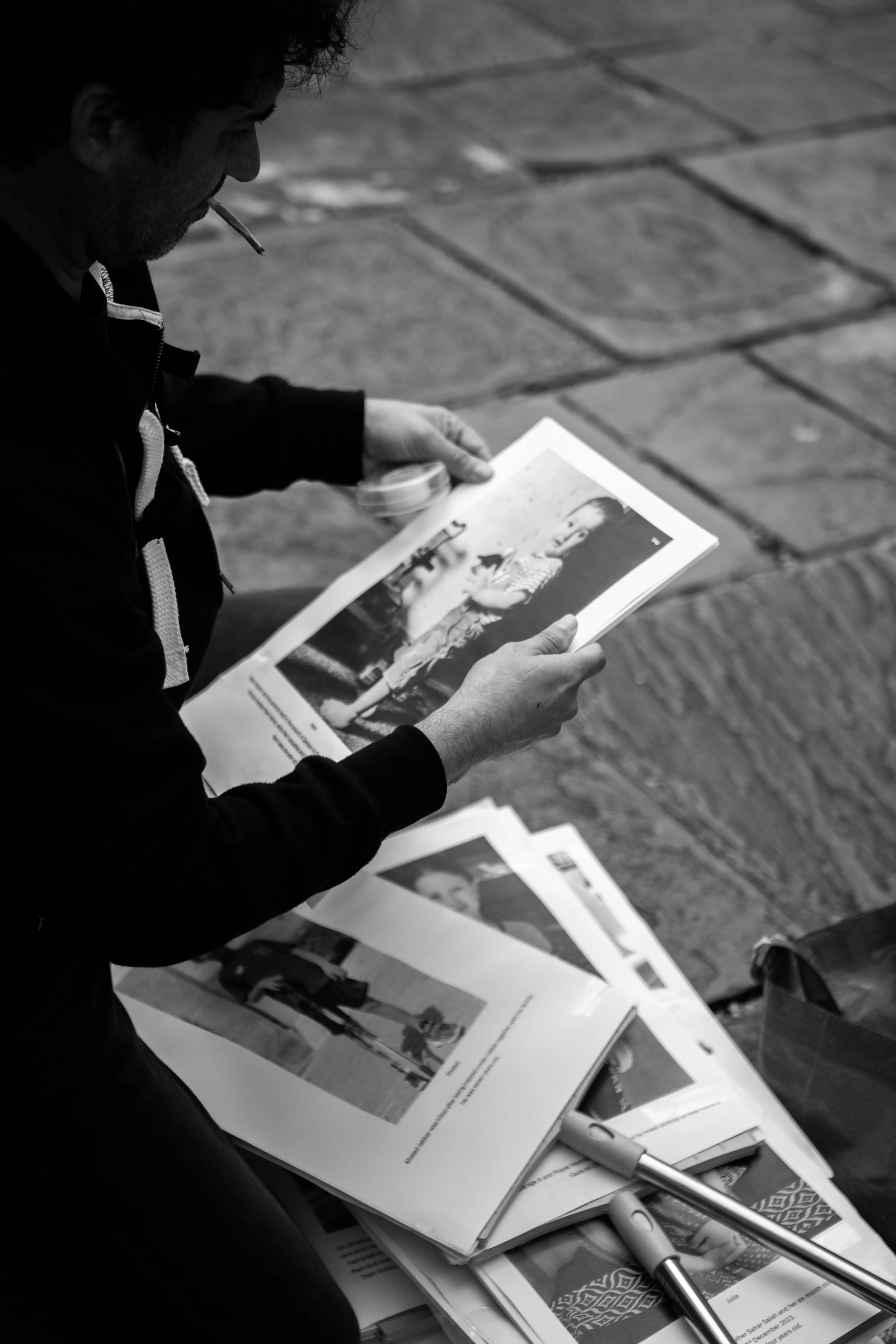 A person with curly hair and a cigarette in their mouth, looking at a photograph of a young girl in striped clothing, among several other photographs and printed documents laid out on a table.