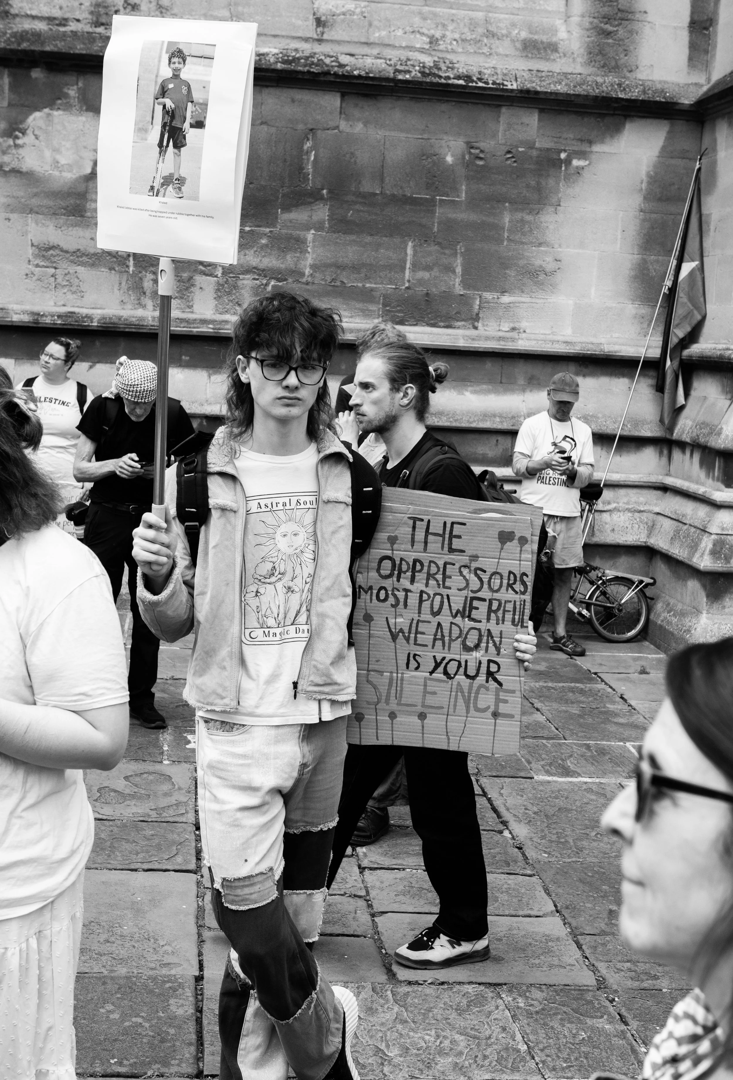 Protesters participating in a demonstration—one person holds a sign with a picture of a young individual with crutches, and another holds a cardboard sign reading, "The oppressors most powerful weapon is your silence." Others in the background are ga