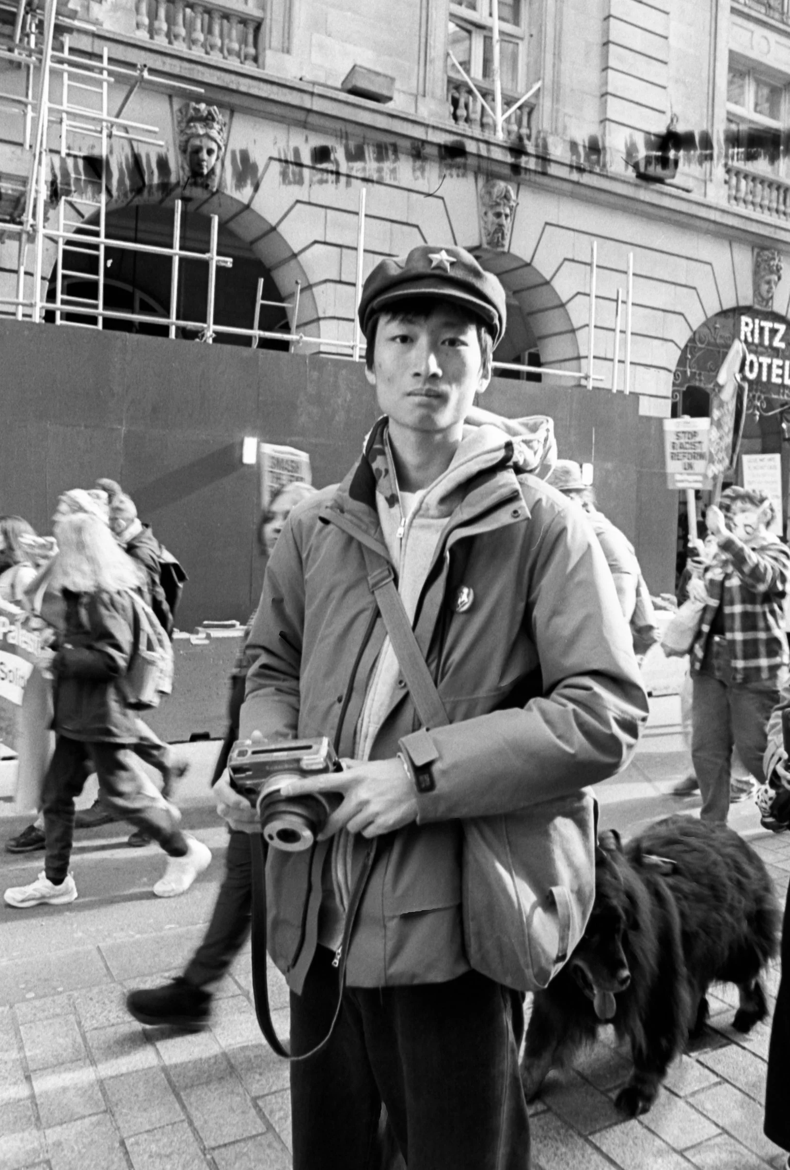 A young person standing in a street with a camera, surrounded by protesting people holding signs, with historical buildings and statues in the background