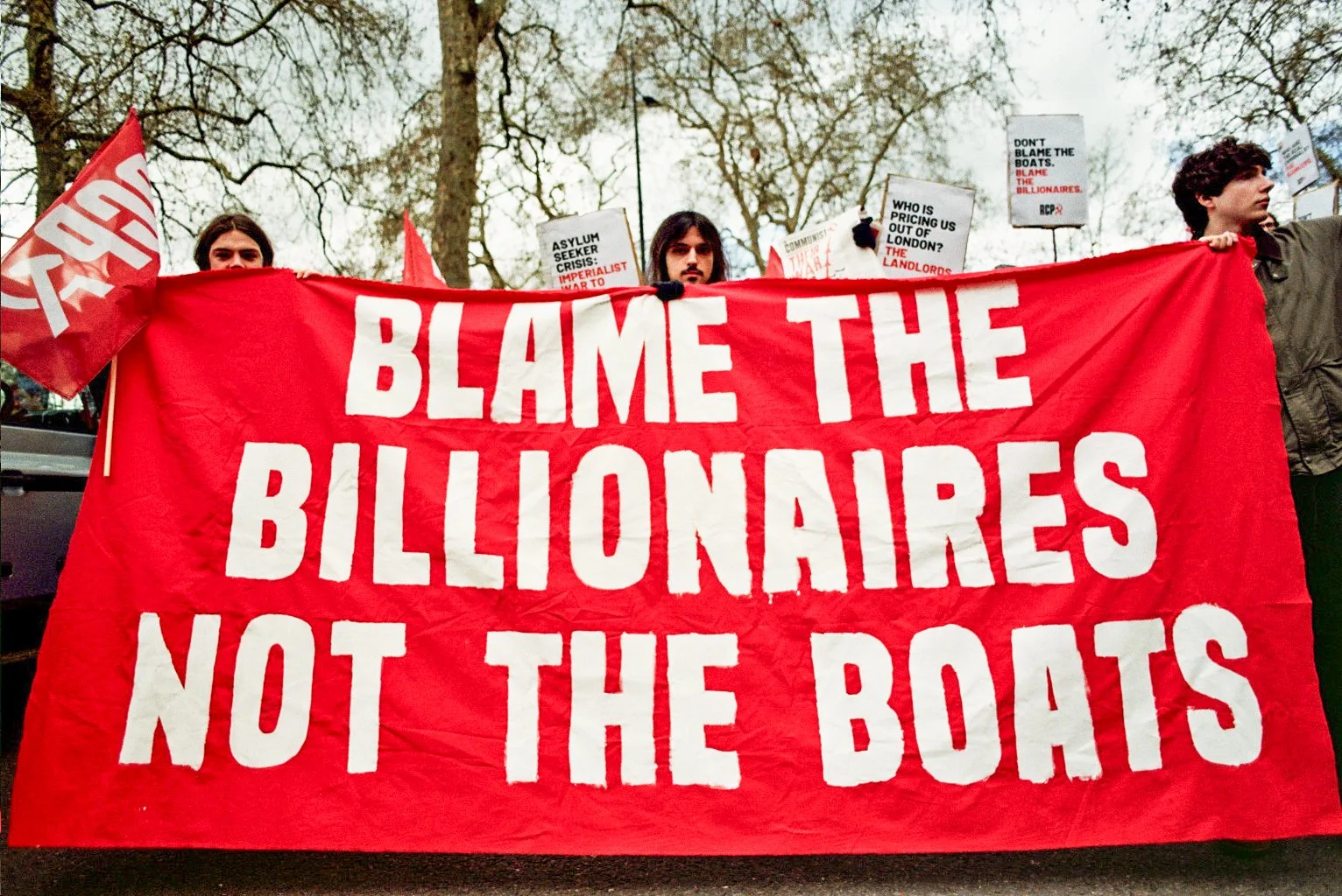 Protesters holding a large red banner that reads 'Blame the Billionaires, Not the Boats' at an outdoor rally with trees in the background.