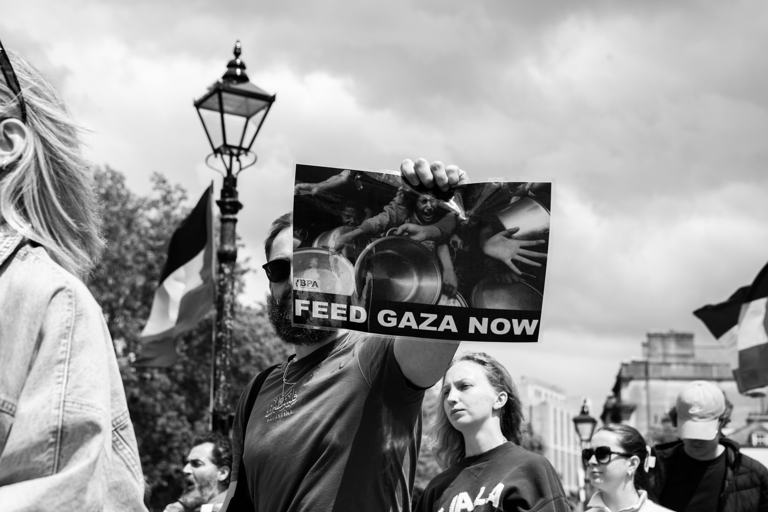 A protester holding a sign that reads 'FEED GAZA NOW' during a demonstration, with other protesters in the background, under a cloudy sky.