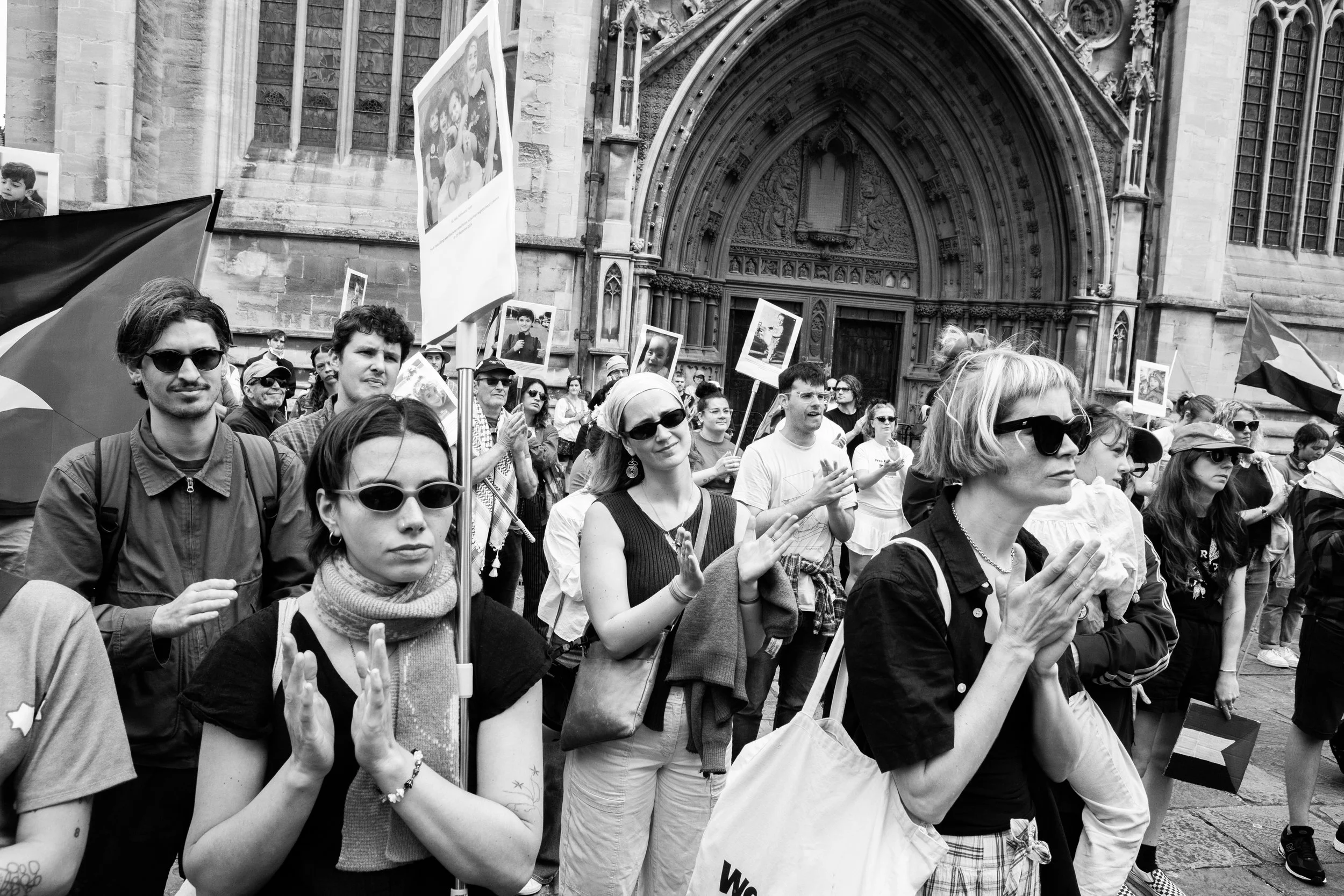 A group of people protesting outside a historic building, some holding signs and flags, some clapping, in black and white.