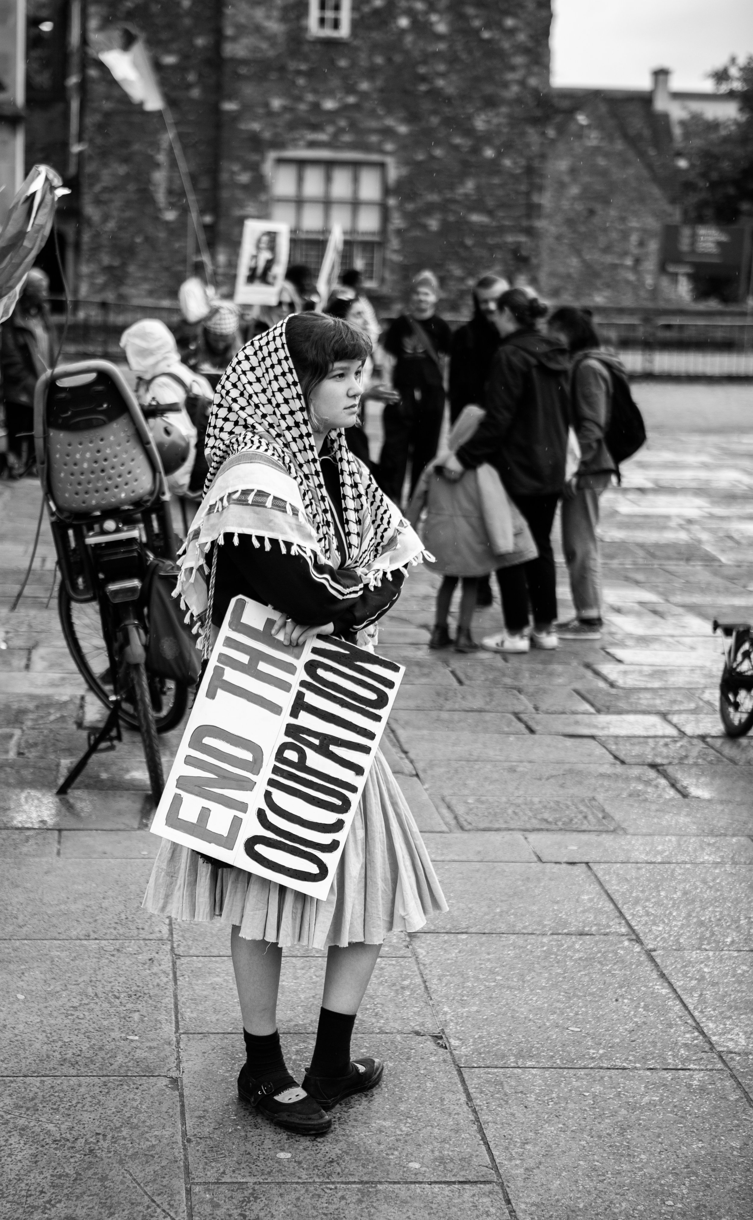 A young girl wearing a patterned headscarf and a pleated skirt holding a sign that says "End the Occupation" during a protest or demonstration on a city sidewalk with a group of people in the background.