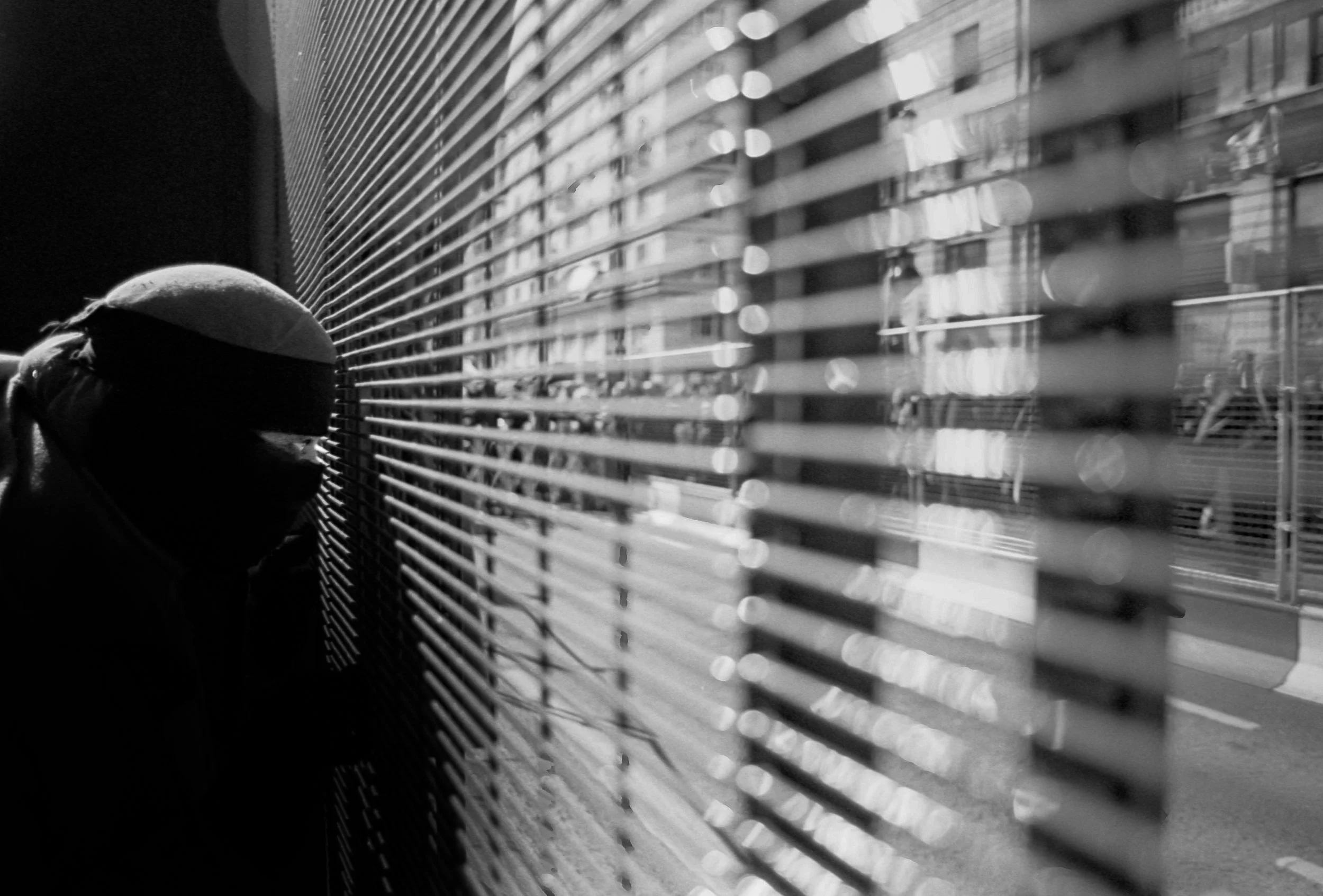 A silhouette of a person wearing a hood and face mask looking through window blinds in black and white.