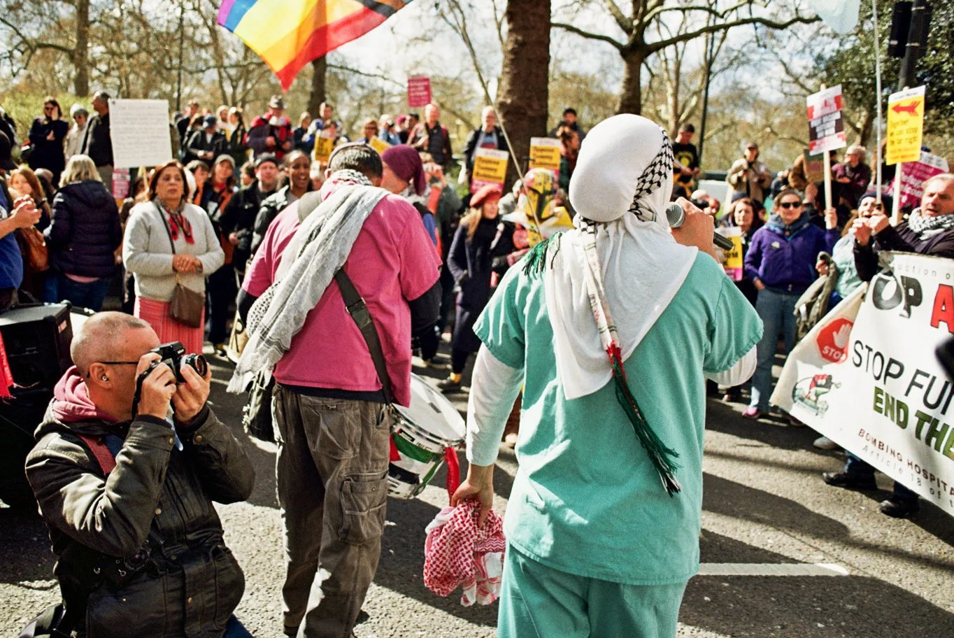 Protest rally with a diverse crowd of people holding signs, banners, and flags, gathering outdoors in a park-like setting during daytime.