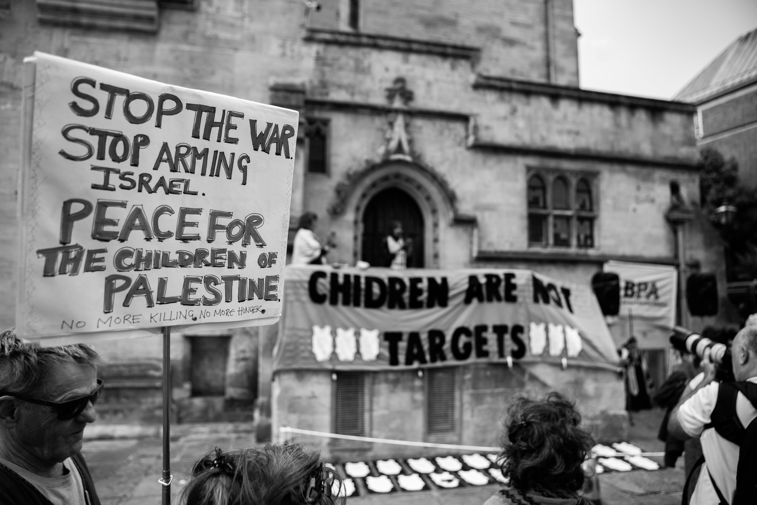 Protest with signs reading "Stop the war, stop arming Israel," and "Children are not targets" outside an old building with people holding cameras.